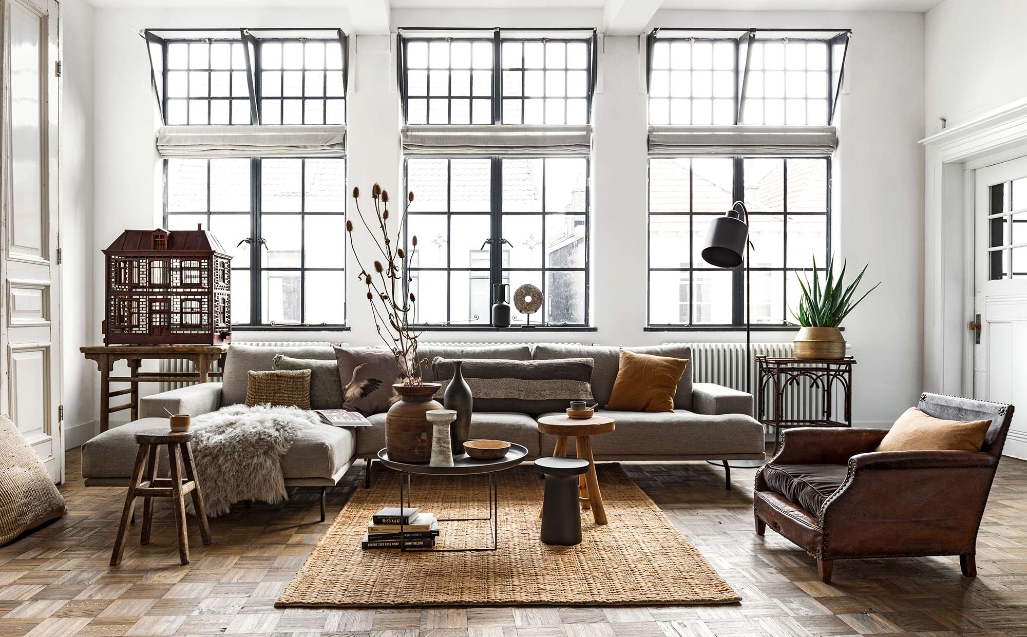 Loungebank Bobbie, ligelement, oud uniek houten tafeltje links naast de bank, zwarte metalen salontafel Norr Duke Large, hoge oude houten kruk, matzwarte vloerlamp Boris, messingkleurige vaas met daarin een aloë vera, ronde steen op voet, diverse kunstboeken, grote houten pot (loftloft.nl). Antieke vogelkooi van begin 1900, antieke leren fauteuil van begin jaren 30, antiek exotisch theetafeltje van begin jaren 30, rechts naast de bank (interieur-vivre.nl). Houten kruk Modern Earth, zwarte houten kruk Shizu, jute vloerkleed van BePureHome, Mongools schapenvacht van Lene Bjerre, kussen met vogels Aza van Claudi Design, camelkleurige kussens van By Mölle, langwerpige wengé houten carved vaas van Scapa Home, zwart kruikje van keramiek (vtwonen.nl). Zeegras kussentje Mekong Nights, grijs linnen kussen met franjes Indochine Mornings, linnen langwerpig bruin-crème gestreept kussen Mekong Nights, zeegras kussen Indochine Mornings op de vloer (aaimadewithlove.com). Cappuccinomokje Basalt Grey, mokje Walnut, lepeltjes, schaaltje Walnut (annemiekebootsceramics.nl). Bamboe schaaltje en wit vintage vaasje privébezit.