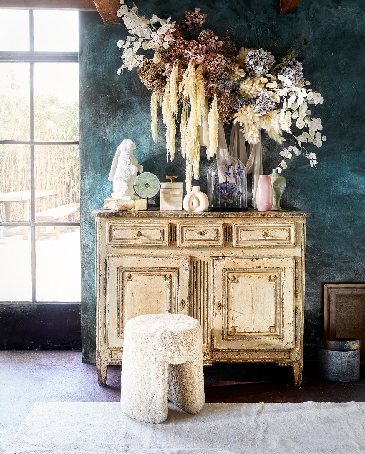 Antieke kast pietjonker.nl). Handgemaakt bloemstuk prijs op aanvraag (judithslagter.nl). Muurverf van Via eStudio prijs op aanvraag, vloerverf Marron van Carte Colori, steen, stolp met vlinders, lijst (brechtmurreatelier.nl). Kelim Turkije, 310×220 cm(couleurlocale.nl). Witte kandelaar Complot van Hay, 13,5×14,5 cm (Øxh) (vtwonen.nl). Kruk met vacht Sequoia, groen rond stenen object, 15 cm (h) (badenbaden.nl). Object konijn (thewunderkammer.eu). Hoge witte doos met hoorn erop, groene vaas (luxurybynature.nl). Roze glazen vaas (hellemardahl.com). Lage doos met marmerprint (maisonnl.com). Doos rechts op vloer prijs op aanvraag (com-anders.nl). vtwonen 06-2021 | styling Liza Wassenaar | fotografie Jeroen van der Spek