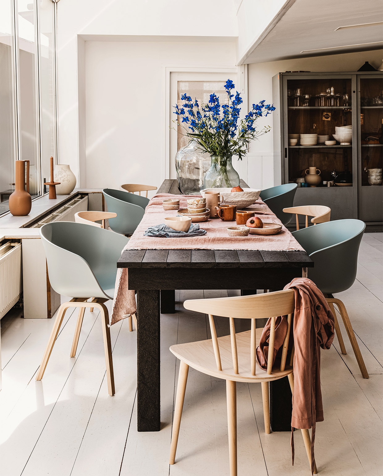 Tafels Standard Long Tables, gerecycled plastic en staal, ook geschikt voor buiten, 60x180x76 cm (bxlxh) per stuk € 1876,- (inekehans.com). Lichtblauwe eetkamerstoelen About a Chair AAC22 van Hay, per stuk € 279,-, beige houten eetkamerstoelen J104 van Hay, per stuk € 255,- (vtwonen.nl). Flesvaas Recycled, 56 cm (h) € 49,95, vaas Evita transparant large € 29,95, witte papier-maché schaal € 49,95, terra mok Svelte € 12,95, roze kom Good Morning € 13,95 (loods5.nl). Roze schaaltje Home Chefs Ceramics € 7,55, roze diep bord Home Chefs Ceramics € 15,95 (hkliving.nl). Roze linnen tafelkleed in kleur Blush, 145×270 cm € 149,-(bymolle.com). Lichtblauwe linnen servetten van H&amp;M Home, set van 2 € 9,99 (hm.com). Roze blouse € 29,95 (zara.com). Terra kandelaar op vensterbank, vanaf € 5,99 (karwei.nl). Overig privébezit. JA | vtwonen 06-2022 | styling Moniek Visser | fotografie Denise Pronk