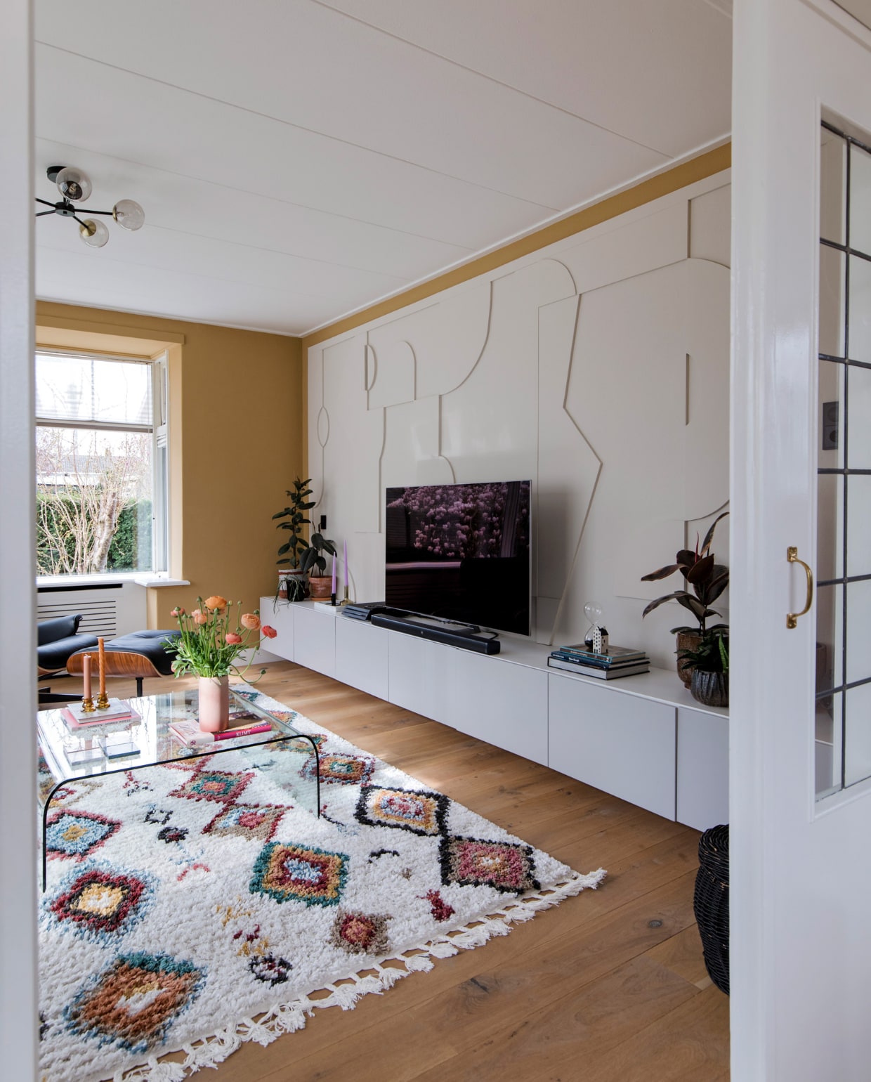 Salontafel Fiam, vloerkleed Sezze van Trendcarpet, Eames Lounge Chair en Ottoman van Vitra. Plafondlamp Globe van Made. Vaas kringloopwinkel, tv-meubel Ikea, 3D-wand zelfgemaakt, tv Samsung.Vloer wit geolied eikenhouten parket via Anderz, radiatorbekleding Cando, raambekleding Verasol. Muurverf Ginger Gold Le Noir et Blanc en Skimming Stone Farrow &amp; Ball. vtwonen 12-2022 | fotografie Corine Hoge Bavel