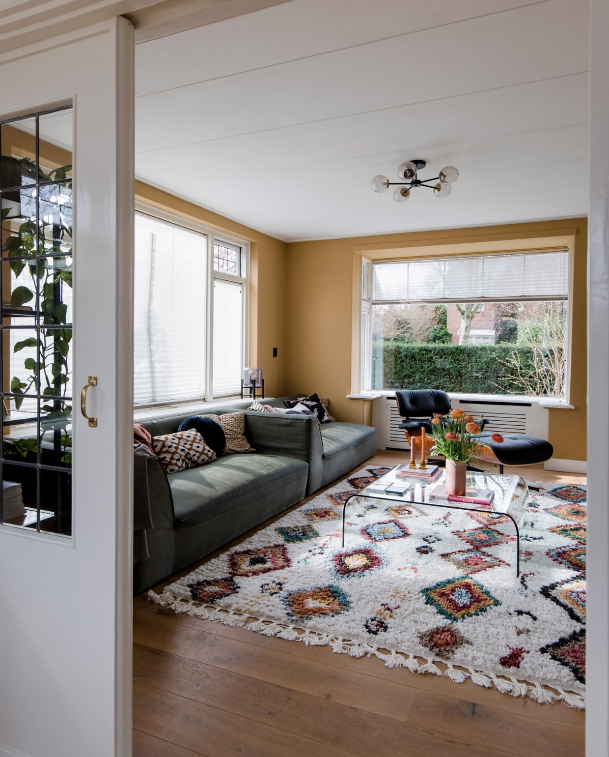 Glazen salontafel Fiam, vloerkleed Sezze van Trendcarpet, plafondlamp Globe van Made, Eames Lounge Chair in palissander van Vitra, vaas uit kringloopwinkel, kussens van Karwei, HKliving en Dutch Decor, kandelaars Kwantum, vloer wit geolied eikenhouten parket via &amp;Anderz, radiatorbekleding Cando, raambekleding Verasol. Muurverf Ginger Gold Le Noir et Blanc. Overig onbekend. vtwonen 12-2022 | fotografie Corine Hoge Bavel