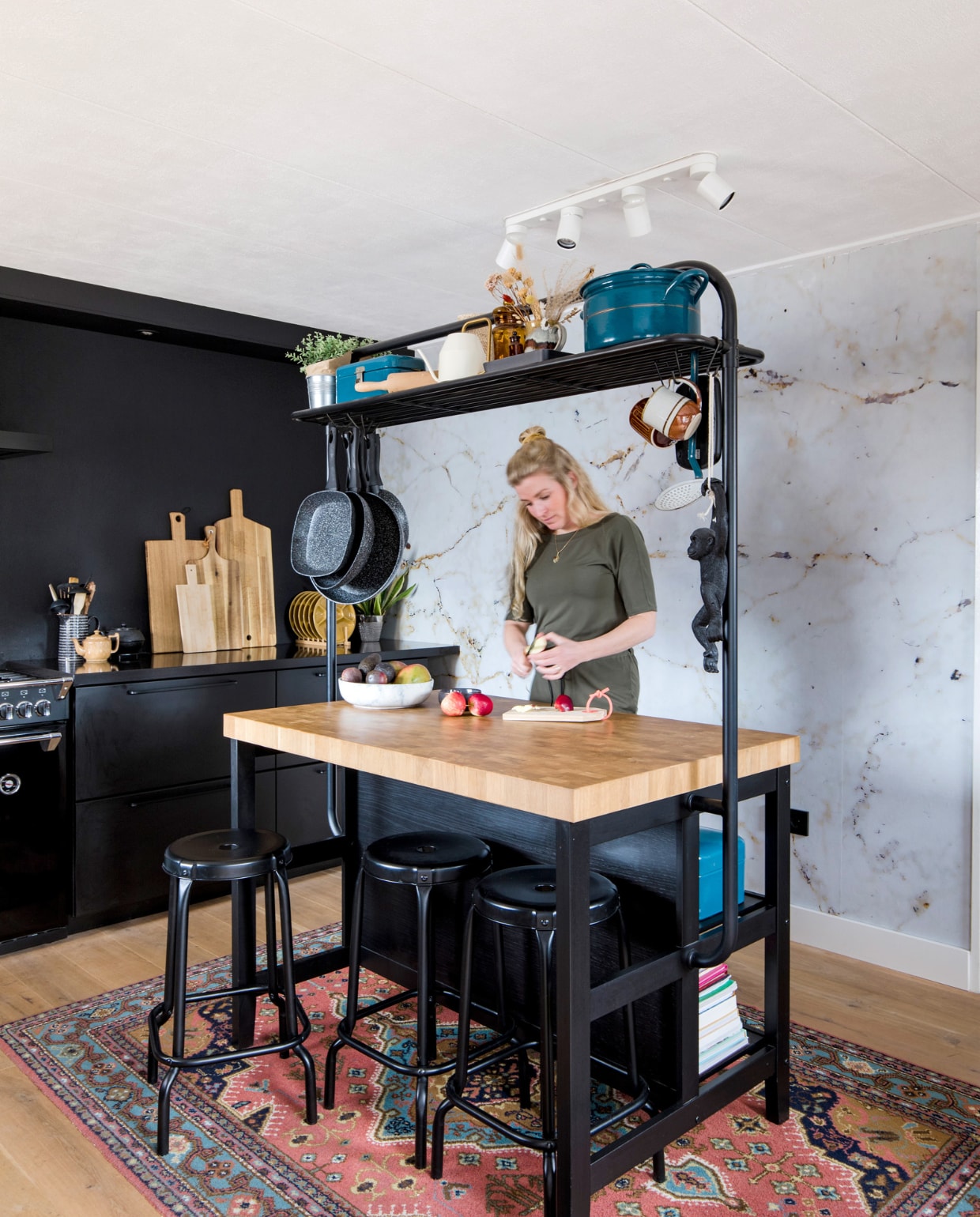 Keuken, hoge tafel, krukken en plafondlamp Ikea. Fornuis en afzuigkap Stoves. Karpet, blauwe emaillen pan, beschuit- en broodtrommel familiestukken. Marmerbehang Graham &amp; Brown. Bruine theepot, hangende seventies mokken, apothekerspotten en okergele borden kringloopwinkel. Zwarte theepot Bredemeijer. Snijplanken van Broodt, Xenos, Hema en Ikea. Fruitschaal Bowls and Dishes, hangende pannen Blokker. Muurverf Off Black Farrow &amp; Ball. vtwonen 12-2022 | fotografie Corine Hoge Bavel