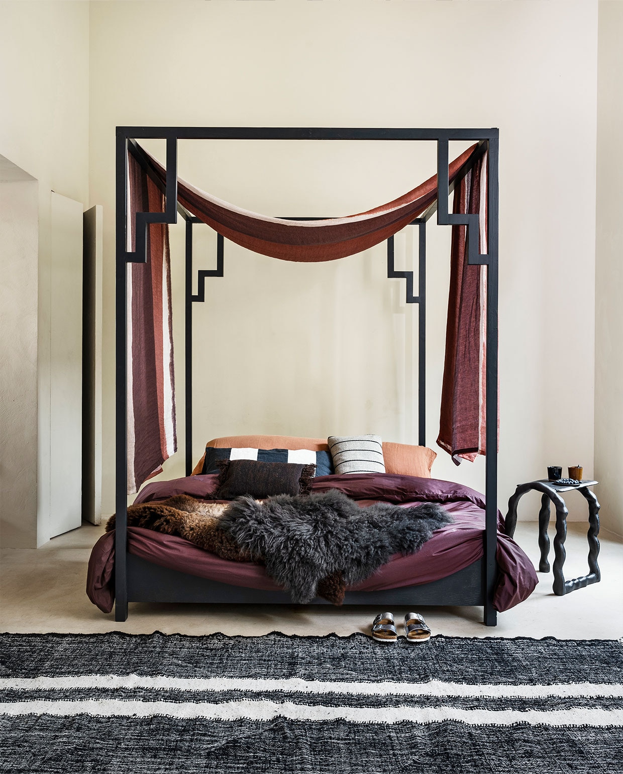 Hemelbed Oriënt, ontwerp Jeanette Spaansen, prijs op aanvraag (com-anders.nl). Linnen hemelstof Kanoko van Osborne &amp; Little, 306 cm breed, per meter € 126,- (wva.nl). Schapenvachten vanaf € 100,- (vanburenbolsward.nl). Stalen bijzettafel van Ferm Living, 45x32x54 cm (bxdxh) € 505,-, gestreept kussen van Ferm Living € 55,-, bruin kussen van Ferm Living € 55,- (misterdesign.nl). Mokken van Serax, per stuk € 10,50 (vtwonen.nl). Wollen vloerkleed, 300×120 cm € 289,-, langwerpige zwart-witte kussenhoes € 69,90 (10dayslifestyle.nl). Katoenen dekbedovertrek € 79,95, extra lang hoofd­kussen Bedmate 160×50 cm € 89,95, linnen sloop terra € 34,95 (suite702.com). Schoenen (birkenstock.com). vtwonen 10-2021 | styling Marianne Luning | fotografie Stan Koolen