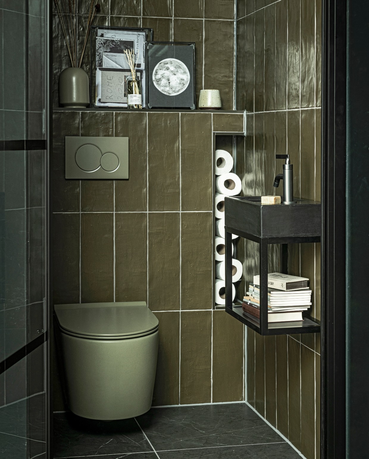 Fontein Tank in kleur Concrete Black, 40x22x10 cm(bxdxh) (ook in Concrete en in White, hoogte 5,5 cm), metalen onderstel Fontein Frame in Zwart,40x22x30 cm (bxdxh), hendel wastafelkraan Grip in kleur Black Steel (ook in Charcoal, Bronze, Brushed en Chrome en met twee hendels), wandtegel Mediterranea in kleur mat Camouflage, 3,2×40 cm, vloertegel Classic Black mat, 74×74 cm (ook in 74×148, 30×60 en 7,3×30 cm). M.m.v. tegeldepot (toilet), ikea.nl (vaas), vtwonen.nl (fotolijsten). vtwonen collectie 08-2020 badkamer | styling Kim van Rossenberg | fotografie Sjoerd Eickmans