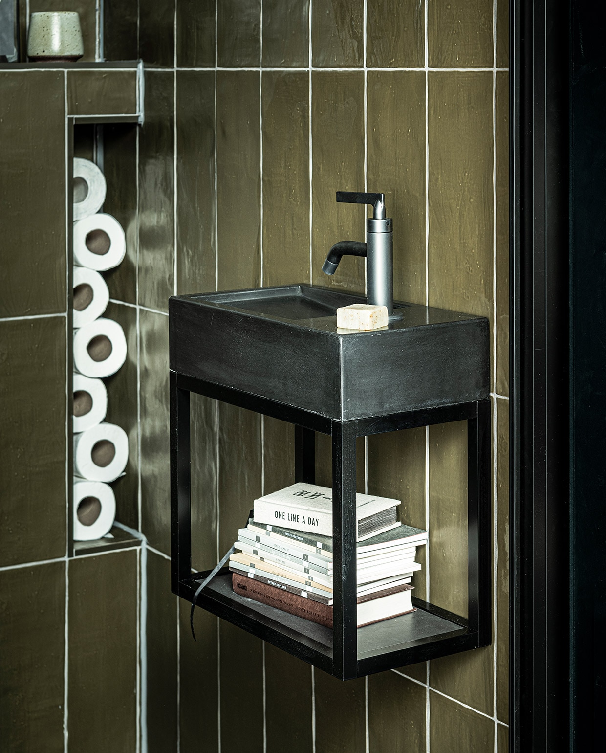Fontein Tank in kleur Concrete Black, 40x22x10 cm (bxdxh) € 299,- (ook in Concrete en in White, hoogte 5,5 cm), metalen onderstel Fontein Frame in Zwart, 40x22x30 cm (bxdxh) € 339,-, hendel wastafelkraan Grip in kleur Black Steel € 459,- (ook in Charcoal, Bronze, Brushed en Chrome en met twee hendels), wandtegel Mediterranea in kleur mat Camouflage, 3,2×40 cm per m² € 61,75, vloertegel Classic Black mat, 74×74 cm per m² € 73,05 (ook in 74×148, 30×60 en 7,3×30 cm).WC uit vtwonen collectie: tegeldepot (toilet), ikea.nl (vaas), vtwonen.nl (fotolijsten). vtwonen 08-2020 | styling Kim van Rossenberg  | fotografie Sjoerd Eickmans
