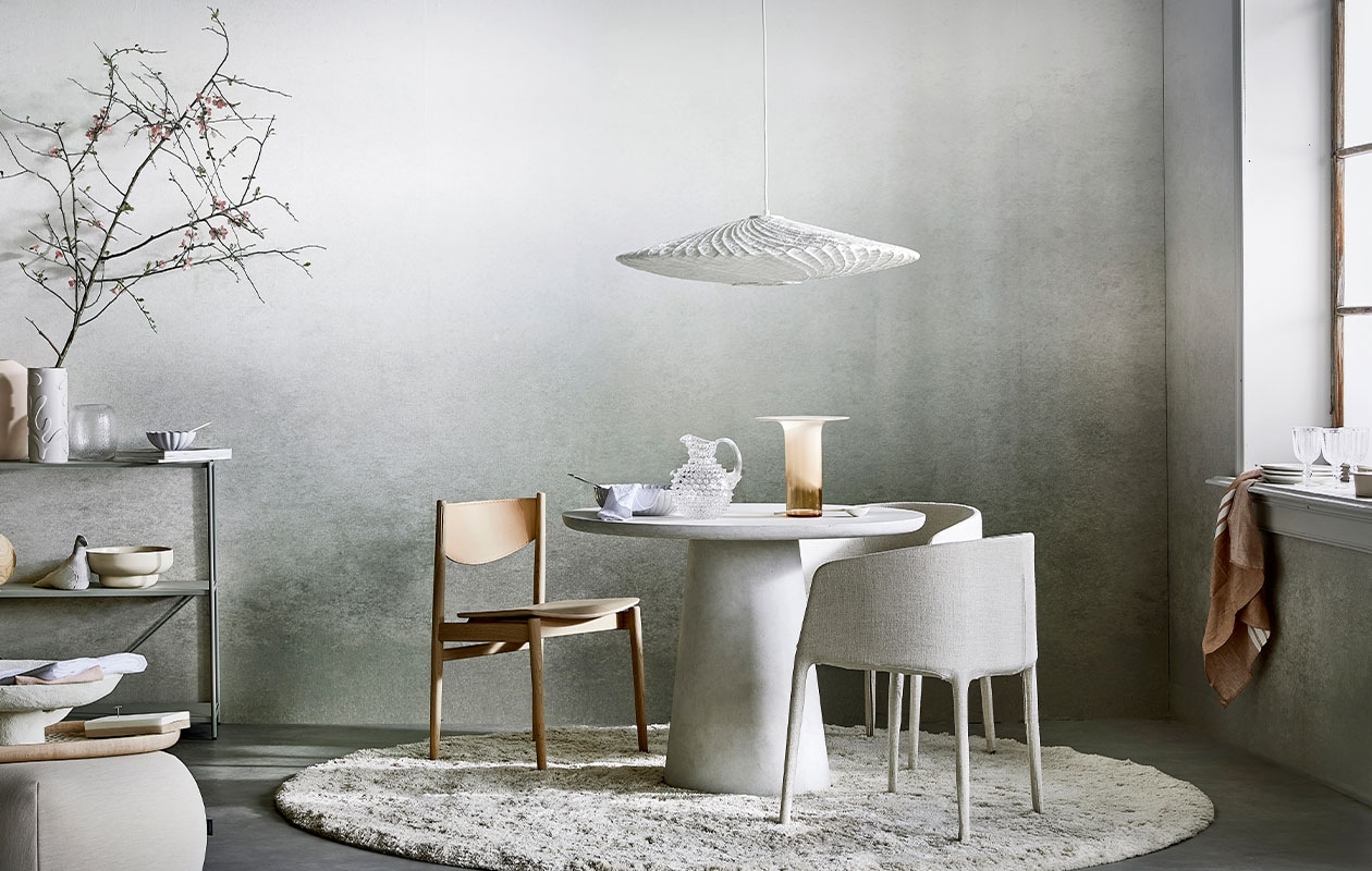 Digitaal behang Degrade Warm Grey, 6 banen van 280×50 cm, totaal (lxb) 280×300 cm. M.m.v. loods5.nl (stellingkast), loods5.nl/design (roze vaas, witte papier-maché schaal, roze schaal), hm.com (witte vaas), bolia.com (glazen vaas, gebroken witte schaal, houten stoel met leren rugleuning), paviljoennoord.com (witte geschulpte schaal klein en groot, wit servet, lepel, glazen kan, roze theedoek, witte borden, wijnglazen), homestede.nl (stoffen vogel, houten dienblad, notitieblok, glazen vaas in koraalkleur), kasstoor.nl (poef, vloerkleed, gestoffeerde eetkamerstoelen, rond vloerkleed), hkliving.nl (hanglamp), basiclabel.nl (eetttafel Damon), letoile.store (witte porseleinen lepel, theedoek), pure-original.nl (lakverf in kleur Ashes). Overig privébezit. vtwonen 05-2021 | productie Tanja Saarberg | styling Danielle Verheul | fotografie Peggy Janssen | assistentie Niels Kist, Danique van Rijn