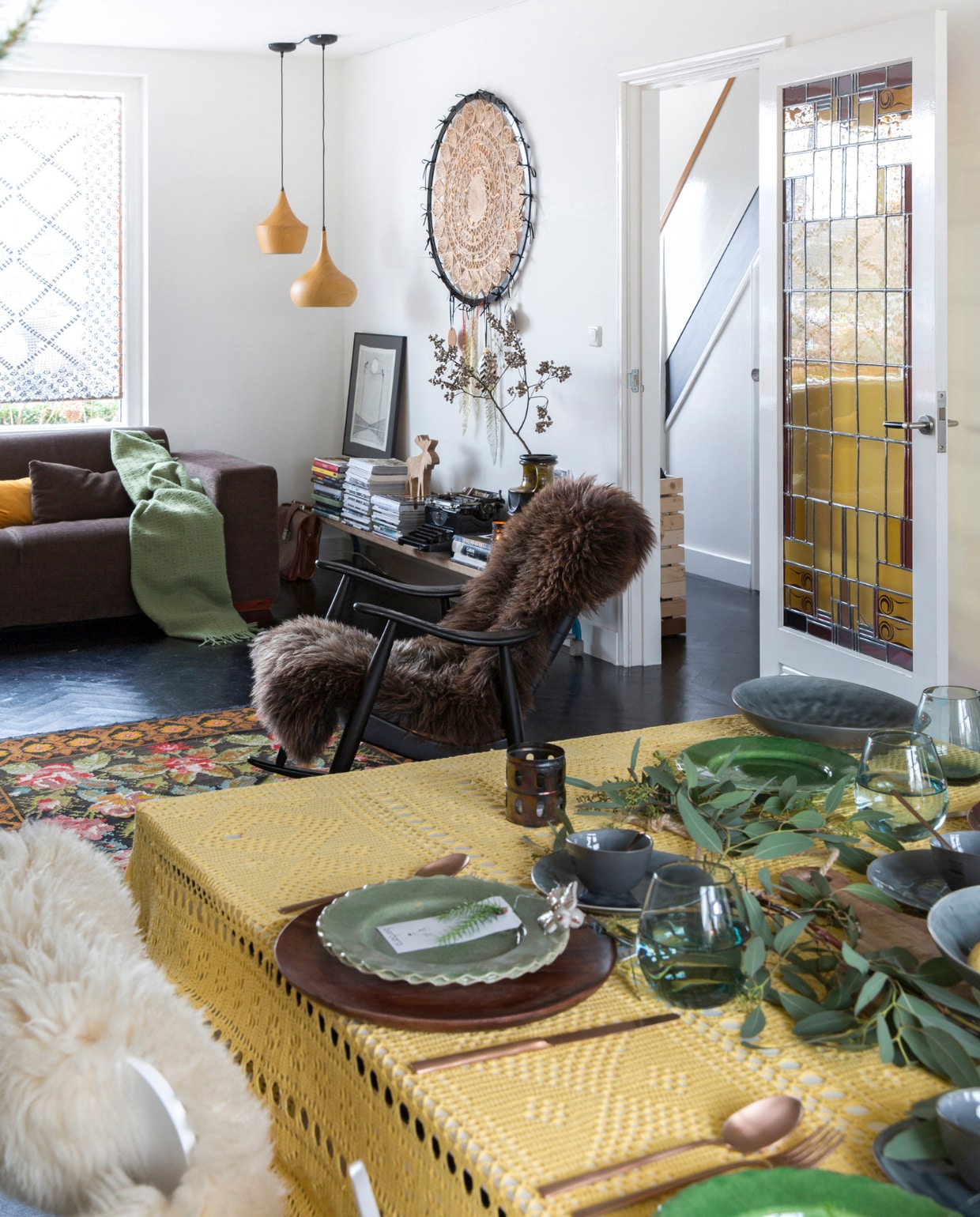 Bank Montèl, plaid Stelling Wonen, geel kussen H&amp;M Home, bruin kussen zelfgemaakt. Raambekleding zelfgemaakt. Schommelstoel grofvuil, bekleding Kay Kesting, vacht kerstmarkt De Rijp. Vloerkleed uit Moldavië. Glas-in-looddeur antiekboerderij in Krommenie. Plafondlampen Loods 5. Dromenvanger zelfgemaakt. Sidetable oude gymbank van de sportschool. Op sidetable v.l.n.r. schilderij uit Portugal, tijdschriften, houten hert Stelling Wonen, vintage typemachine, boeken, vaas Berlijnse rommelmarkt. Servies, glaswerk, bestek, kaars met ster, groene keramieken kan K’ook!. Vlinderstoel Fritz Hansen, witte vacht Ikea. Groene takken Floribundus. Visgraatvloer Van Dijk &amp; Ko, zwart geschilderd en gelakt met producten van Sigma, muurverf RAL 9010 en matzwarte muurverf. vtwonen 13-2022 | styling Barbara Natzijl | fotografie Margriet Hoekstra