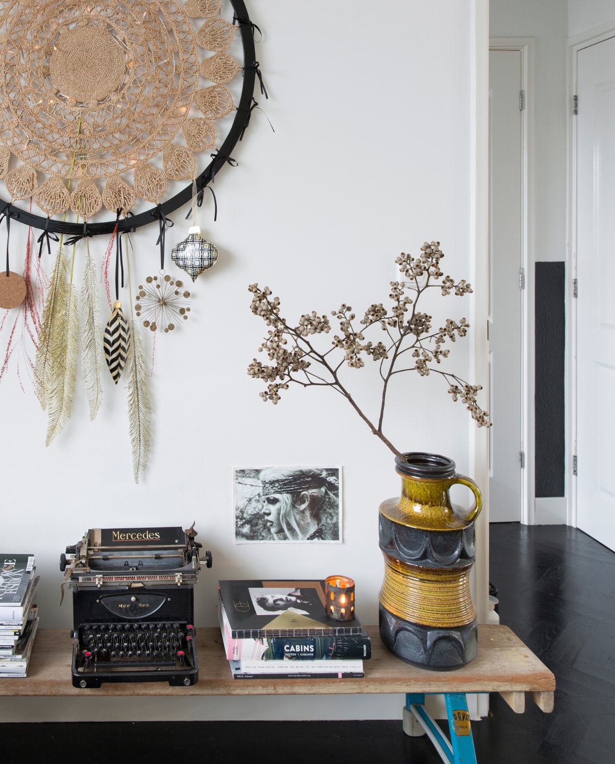 Dromenvanger zelfgemaakt. Foto Brigitte Bardot Goed in Stijl. Sidetable oude gymbank van de sportschool. V.l.n.r. tijdschriften Vogue,vtwonen, Elle Decoration, Soffa Magazine, typemachine Mercedes gevonden in container, mode-,interieur- en reisboeken, kaarsenhouder Stelling Wonen, vaas Berlijnse rommelmarkt, tak Floribundus. Visgraatvloer Van Dijk &amp; Ko, zelf zwart geschilderd en gelakt met producten van Sigma, muurverf RAL 9010 en matzwarte muurverf.  vtwonen 13-2022 | styling Barbara Natzijl | fotografie Margriet Hoekstra | tekst Sunna Borghuis