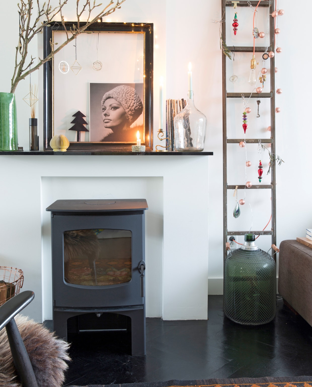 Kachel Deyle. Op de schoorsteenmantel v.l.n.r. tak Floribundus, lijst gekregen, lichtjes om lijst Karwei, geurstokjes Frau Tonis Parfum, geel papieren bolletje zelfgemaakt, kerstboomkaars K’ook!, foto Sophia Loren Goed in Stijl, kaarsenstandaards en vaas rommelmarkt, oude lp’s. Messing ladder uit oude torpedojager van een afgedankt schip, kerstdecoratie De Glazen Pauw, lichtsnoer Praxis, oude benzinefles rommelmarkt. Vloerkleed uit Moldavië. Visgraatvloer Van Dijk &amp; Ko, zwart geschilderd en gelakt met producten van Sigma, muurverf RAL 9010. vtwonen 13-2022 | styling Barbara Natzijl | fotografie Margriet Hoekstra | tekst Sunna Borghuis