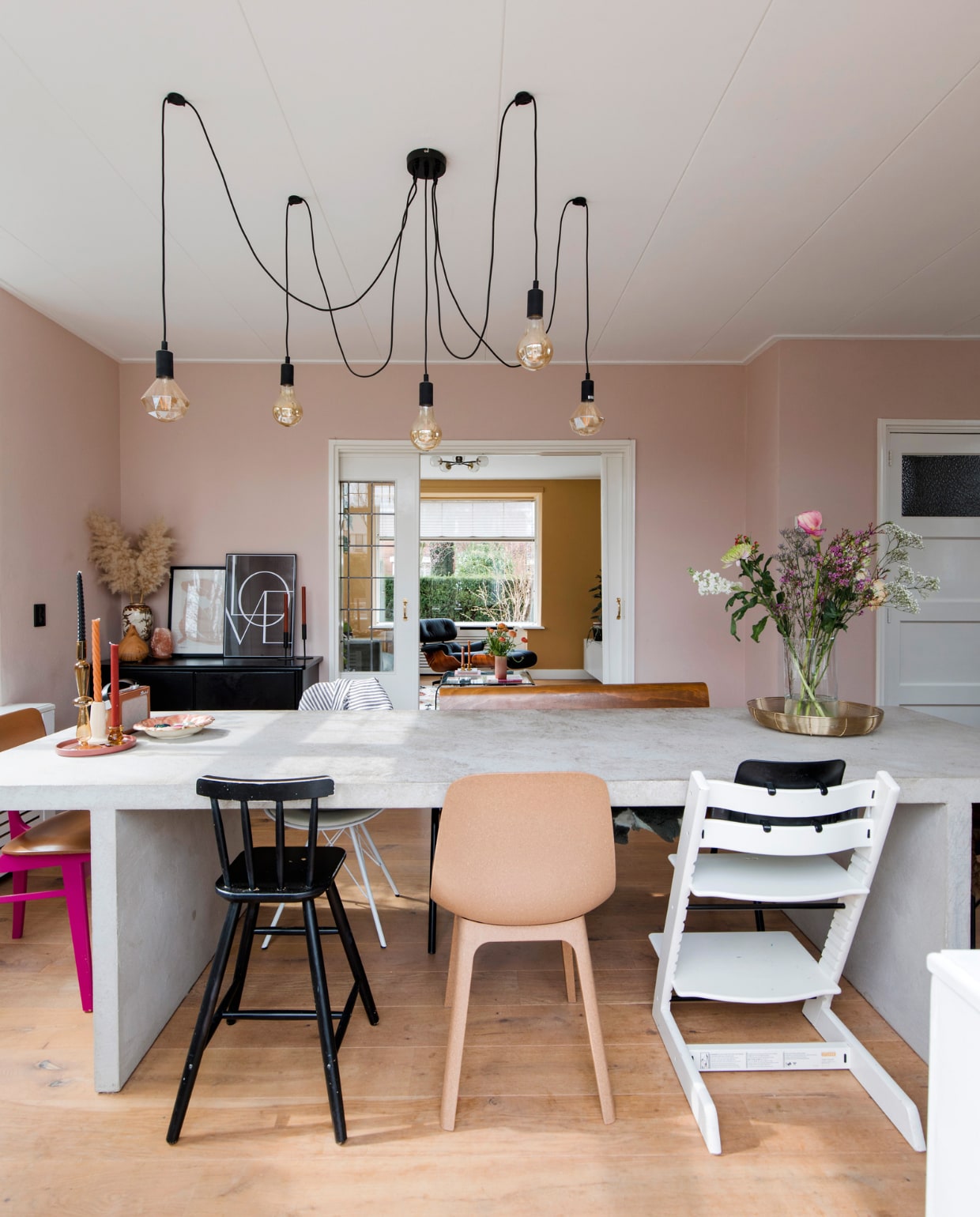 Tafel zelfgemaakt met beton ciré. Felroze stoel vintage. Zwarte kinderstoel, stoel Odger en lijsten Ikea. Witte kinderstoel Tripp Trapp van Stokke. Hanglamp Eglo. Eettafelbank vtwonen. Goudkleurige draadschaal Xenos, kandelaars Kwantum, vintage aardewerken schaal Flora Gouda, Chinese porseleinen vaas familiestuk, posters I am Fy. Muurverf Pink Nudity Flexa. Wit geolied eikenhouten parket &amp;Anderz. Overig onbekend. bk vtwonen 12 2022 | fotografie Corine Hoge Bavel