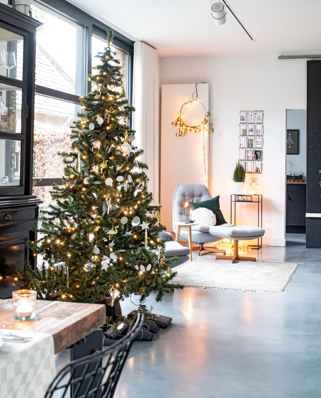 Vintage stoelen Bestwelhip. Groene kussen vtwonen, witte kussen Sissy-Boy. Bijzettafeltje Joelle Boers Studio, stalen bijzettafel tegen de muur Robuust Maatwerk. Kaartenrek Madam Stoltz. Ronde kerstversiering zelfgemaakt. Op de betonnen vloer ligt een kleed van HK Living. vtwonen 12-2019 | Binnenkijken in een vrijstaand huis in Geleen woonkamer kerstboom | styling Babs Dols | fotografie Louis Lemaire/ Insidehomepage