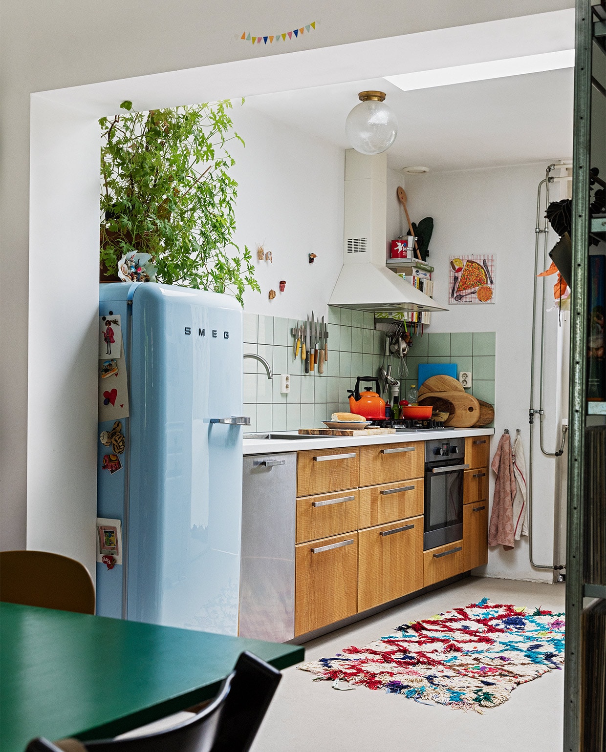 Koelkast Smeg. Geraniumplant zelf gestekt. Kitsch schelp op koelkast souvenir uit Griekenland. Magneet-souvenirs meegenomen van verschillende reizen. Fluitketel en pan Le Creuset. Messen Suncraft, Opinel, Dille &amp; Kamille, Pallarès Solsona. Snijplanken Hay, Ikea, Serax. Soeplepel Brabantia. Rvs keukengerei Dille &amp; Kamille. Schaaltje van een pottenbakkersmarkt. Afzuigkap en oven Ikea. Ovenhandschoen Kook. Pollepel Kookhuis aan de Maes. Handdoeken Sissy-Boy. Tapijt The Souks. Pizza-schilderij Oskar. Ronde plafondlamp lampenwinkel.nl. JA | vtwonen 13-2021 | styling Ilona Jongepier |  fotografie Dana van Leeuwen