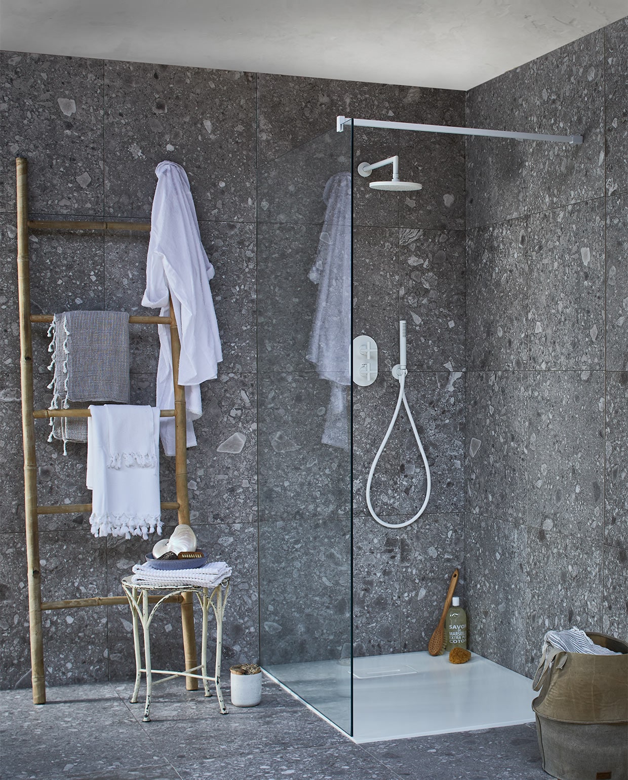 Op de wand en vloer: tegel Composite Dark Grey van vtwonen tegels by Douglas &amp; Jones, douchevloer met geïntegreerde afvoer van gegoten marmer in powder white, glazen douchewand met muurbevestiging in powder white, diverse uitvoeringen (transparant,donkergrijs en spiegelglas), douchekraan Cross, muur-versie powder white met afdekset voor inbouwthermostaat, handdouche, douchearm, hoofddouche. Bamboe ladder, witte badmat op kruk (l’Etoile Conceptstore). Hamamdoeken (Ottomania). Badborstel, nagelborstel (My Deer Store). Witte schaal (Raw Materials). Douchegel, spons (&amp;Klevering). Kruk (Neef Louis). Canvas mand (Hoffz). Schelp (De Weldaad). Pot met kurken deksel (Restored). vtwonen 02-2017 | vtwonen badkamercollectie | styling Cleo Scheulderman | fotografie Jeroen van der Spek | productie Inge Steketee