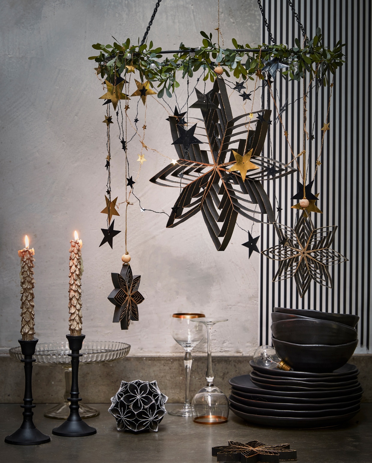 Zwarte metalen hanger, 40×46 cm (Øxh) € 9,99 omhuld met kunststof mistletoe guirlande € 3,99. Aan de mistletoe: diverse zwarte papieren ornamenten, per stuk vanaf € 2,49, goudkleurige metalen slinger met sterren, 210 cm (l) € 3,49 (ook in zilver). Op tafel: goudkleurige kaarsen met glitter, per 2 € 6,99, zwart papieren ornament bloem met glitter, 10 cm (Ø) € 5,99. vtwonen 12-2022 | productie Tanja Saarberg, Isa Hoevens | styling Kim van Rossenberg | fotografie Peggy Janssen