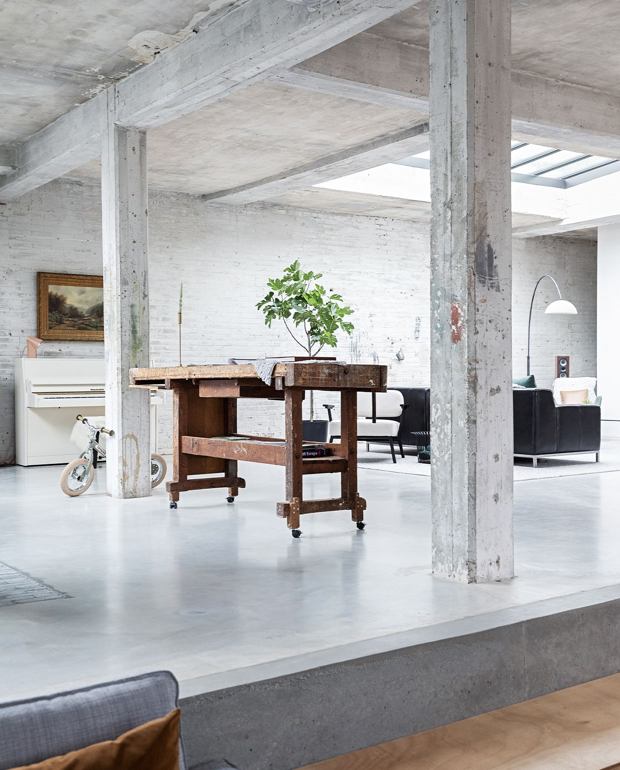 Vloer gevlinderd beton. Hardstenen waaltjes Spotted Bluestone. Werkbank erfstuk meubelfabriek. Kinderfiets Trybike. Witte fauteuil Ode Lounge Chair van Studio Henk.Bank Ikea. Booglamp Venled. Piano en schilderij erfstukken. Lamp op piano Elleke Rutten. vtwonen 01-2022 | styling Ilona Jongepier | fotografie Dana van Leeuwen | visagie Djolien de Kreij