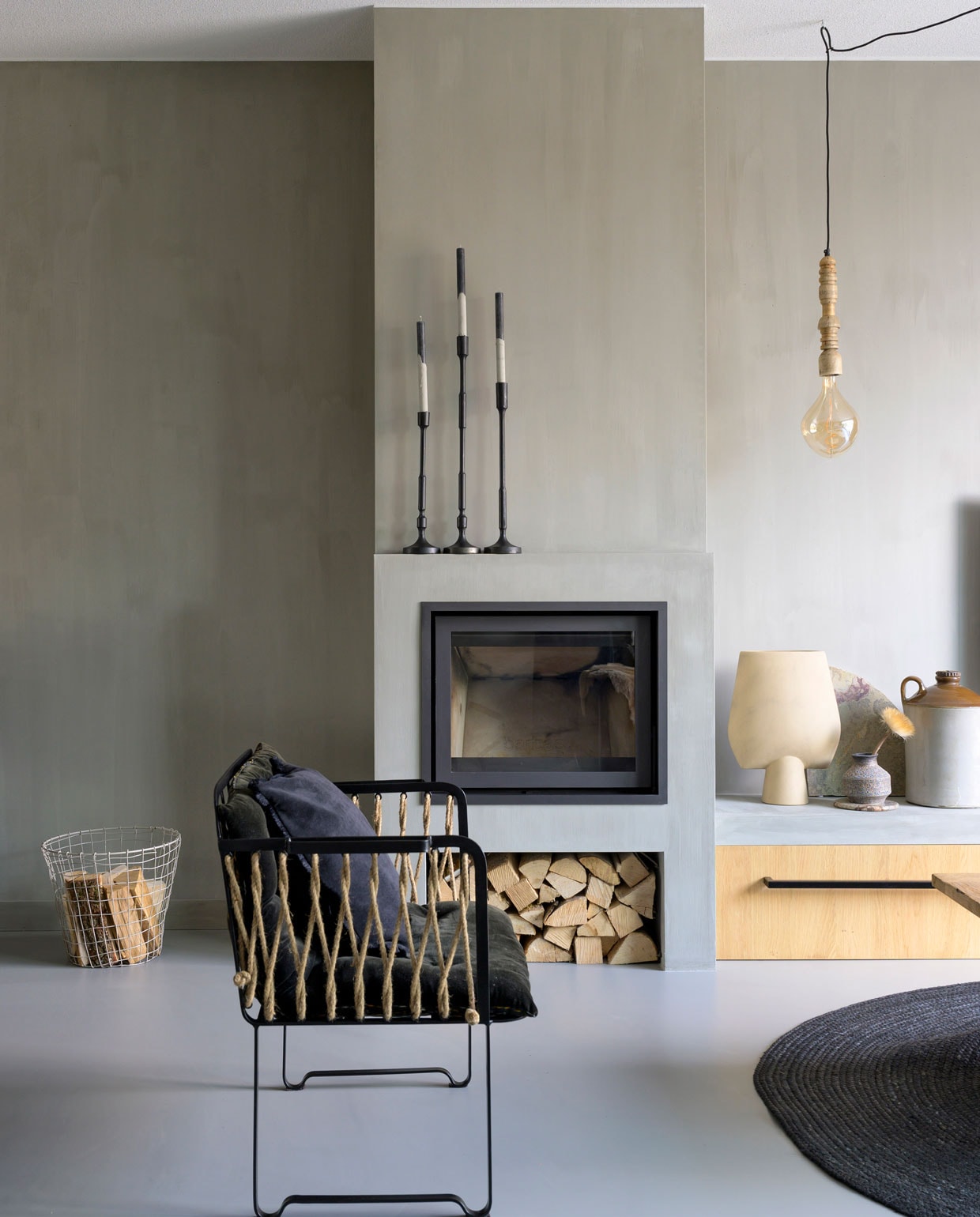 Grijze woonkamer met open haard | Houtkachel Tibas. Wandbank met lade maatwerk. Draadmand Mica Decorations. Kandelaars, hanglamp Casa Vivante. Peertje Hornbach. Vazen Casa Gitane. Grote kruik en fauteuil Madam Stoltz. Vloerkleed Karwei. Muurverf CC020 Zinco van Carte Colori. vtwonen 08-2021: styling José Spaans | fotografie Stephanie Driessen | tekst Christine van der Hoff