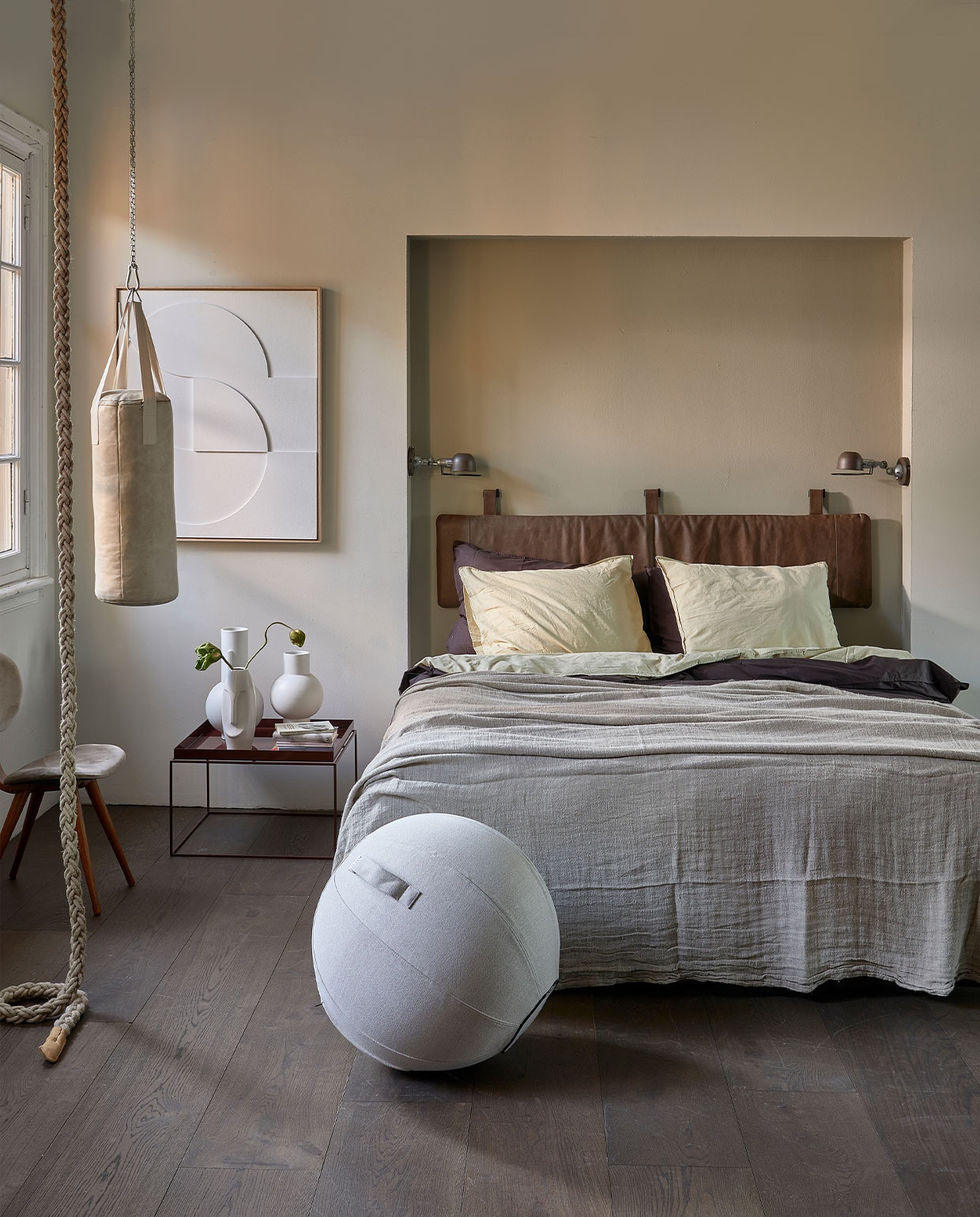 Muurverf krijtlook mat van vtwonen, kleur achter bed Soap, eromheen Linen, per liter € 14,20, vloer laminaat XL, kleur board per m2 € 25,99 (karwei.nl). Hoofdbordkussen met lus en beugel, 40×180 cm € 339,- (nahka.nl). Boxspring Espevar, 180×200 cm € 858,-, dekbed Smasporre, 240×220 cm € 29,99, hoofdkussens Gronamarant, 50×60 cm, per stuk € 19,99, hoeslaken Dvala, 180×200 cm € 29,99, beige/groen katoenen dekbedovertrek en slopen Angslilja, 240×220 cm € 34,99 (ikea.nl). Linnen beddensprei naturel, 270×270 cm € 229,-, matwitte vazen achterste op tafel, 21×32 cm (Øxh) € 42,95, vaas daarnaast 21×42,5 cm (Øxh) € 49,99, relief art paneel € 595,- (hkliving.nl). Veganleren boksbal € 549,- (sukha.nl). Klimtouw € 75,-, stoel Pastoe met fluwelen bekleding € 295,- (neeflouis.nl). Donkerbruin katoenen dekbedovertrek Ambiante, 200×220 cm € 46,25, wandlampen Lucide Honore, per stuk € 55,99, zitbal Vluv Sova, 65 cm (Ø) € 105,-, vaas Erla met gezicht, 31 cm (h) € 24,95, bijzettafel Tray van Hay, kleur chocolate € 228,- (vtwonen.nl). Reliëfkunstpaneel € 625,- (hkliving.nl). vtwonen 05-2022 | Styling Fietje Bruijn | Fotografie Dennis Brandsma\n