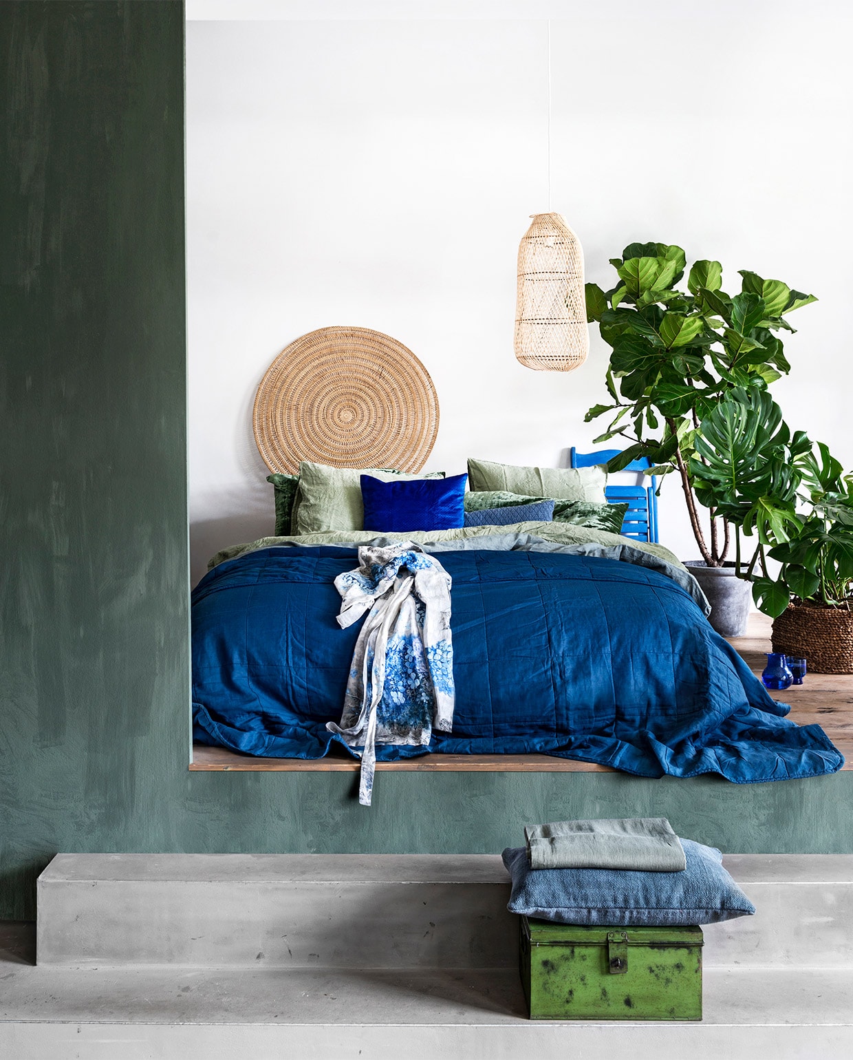 Wand geverfd met kalkverf in kleur Agave per liter € 45,85 (cartecolori.nl). Rotan ornament op muur h€ 69,95, bamboe lantaarn € 88,95, mand van waterhyacinth € 54,95 (manggisliving.nl). Jeansblauwe sprei Gulved, 26×250 cm € 49,95 (ikea.nl). Grijsblauw linnen dekbedovertrek, 240×220 cm € 219,-, linnen kimono Hortensia € 119,-, grijsblauw linnen laken op kist, 240×260 cm € 139,- (timelesslinen.com). Olijfgroen dekbedovertrek, 240×220 cm € 99,-, groen fluwelen kussen, 50×90 cm € 19,99 (hm.com). Hardblauw kussentje Venche € 4,99 (ikea.nl). Grijsblauw linnen/katoen kussen € 49,95, blauwe klapstoel € 29,99, groene metalen kist € 69,95, grijsblauw kussen op kist € 49,95 (hetkabinet.nl). Boompje Lyrata/tabaksplant – vioolbladplant (kwekerij). Plant Monstera Deliciosa plant, betonnen pot (tuincentrum). JA | vtwonen 05-2019 | styling Moniek Visser  | fotografie Stan Koolen