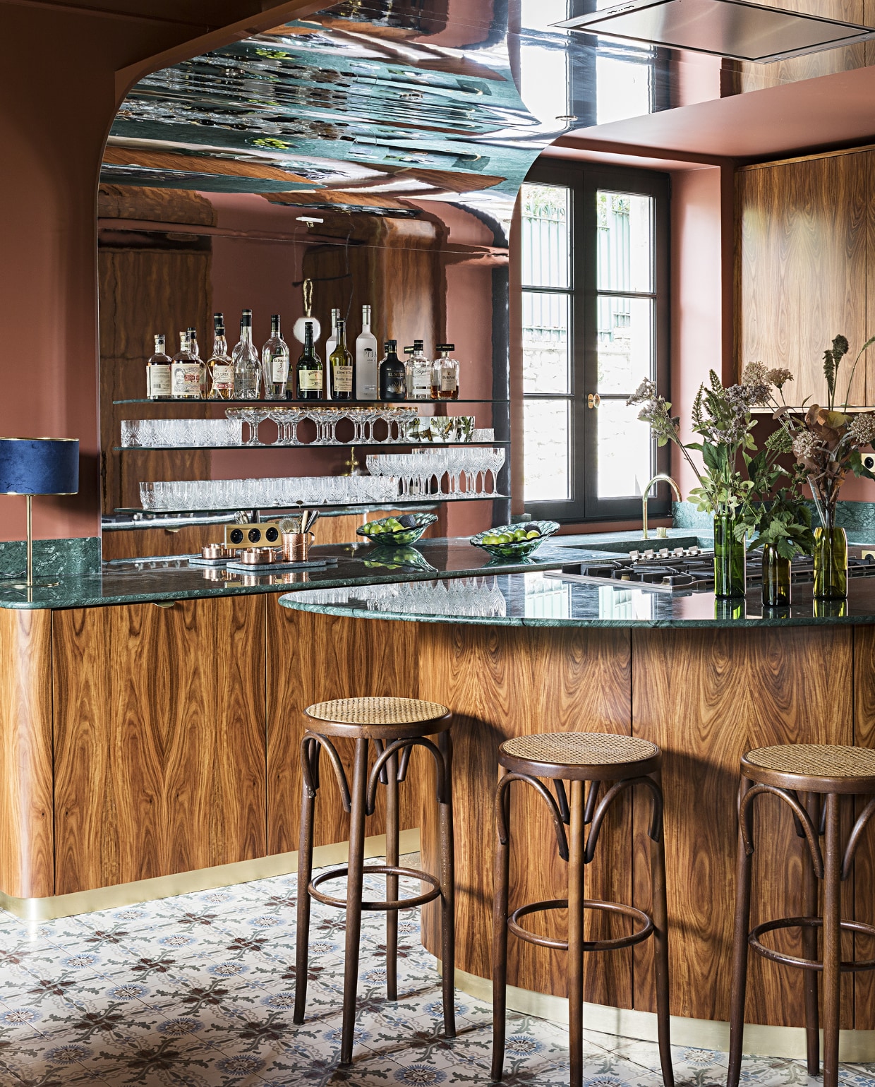 De keuken, ontworpen door Fanny en Fabrice zelf met interieurarchitect Carlos Pujol, is uitgevoerd in palissanderfineer en verde Guatamala (groen marmer). De achterwand is bekleed met spiegelfolie. Muurverf Book Room Red van Farrow &amp; Ball. Barkrukken tweedehands. Overig onbekend. NEE | vtwonen special binnenkijken 2022-3 | Productie AC Scoffoni | Styling Marie-Maud Levron | Fotografie Yann Deret | Tekstbewerking Renate Dahlmans