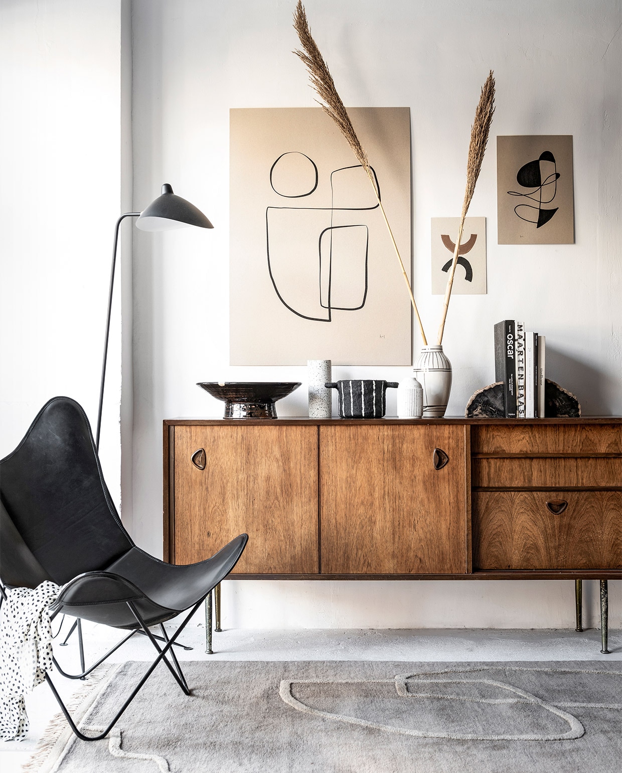 Abstracte inktlijntekening Sola van Henri &amp; Lydia € 6,79, abstracte inkttekening van Henri &amp; Lydia € 6,34, handgemaakte printable art van Miuus Studio set van 2 € 16,17 (etsy.com), alles geprint op grijsbord A4-formaat € 18,15, A3-formaat € 21,18, 70×100 cm € 53,85 (repros.nl). Schaal met lijnenspel € 475,-, lamp Replica Serge Mouille € 349,-, leren vlinderstoel voor Airborne € 999,-, teakhouten dressoir William Watting 19605 € 1295,- (cocooncollectables.nl). Terrazzo vaas van Zakkia € 39,- (restored.nl). Zwarte bloempot van papier € 33,50, wit keramiek vaasje met zwarte spikkels € 13,95 (storewithoutahome.nl). Witte vaas Outline € 26,33 (housedoctor.com). Handgemaakte boekensteunen van versteend hout € 149,- (studiosomee.com). Vloerkleed Desert Tufted Rug Sand, 140×200 cm € 599,- (fermliving.com). Boeken en blouse privébezit. vtwonen 04-2019 | styling Kim van Rossenberg  | fotografie Sjoerd Eickmans