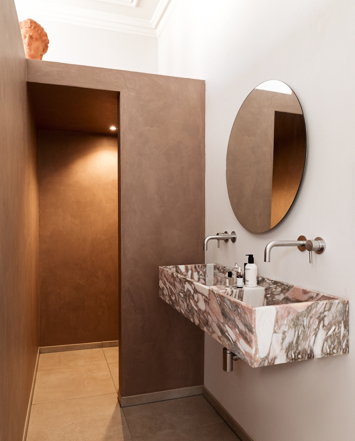 Ronde spiegel badkamerspiegels.nl. Wasbak heeft Louk nagetekend van een foto en laten maken door Swaal Natuursteen. Vloertegels Maxaro. Beton ciré in kleur Caramel Basebeton. RVS-kranen KraanDesign. Handzeep Marie-Stella-Maris. Gezichtsreiniging Image Skin Care. vtwonen 08-2022 | styling Ilona Jongepier |  fotografie Dana van Leeuwen