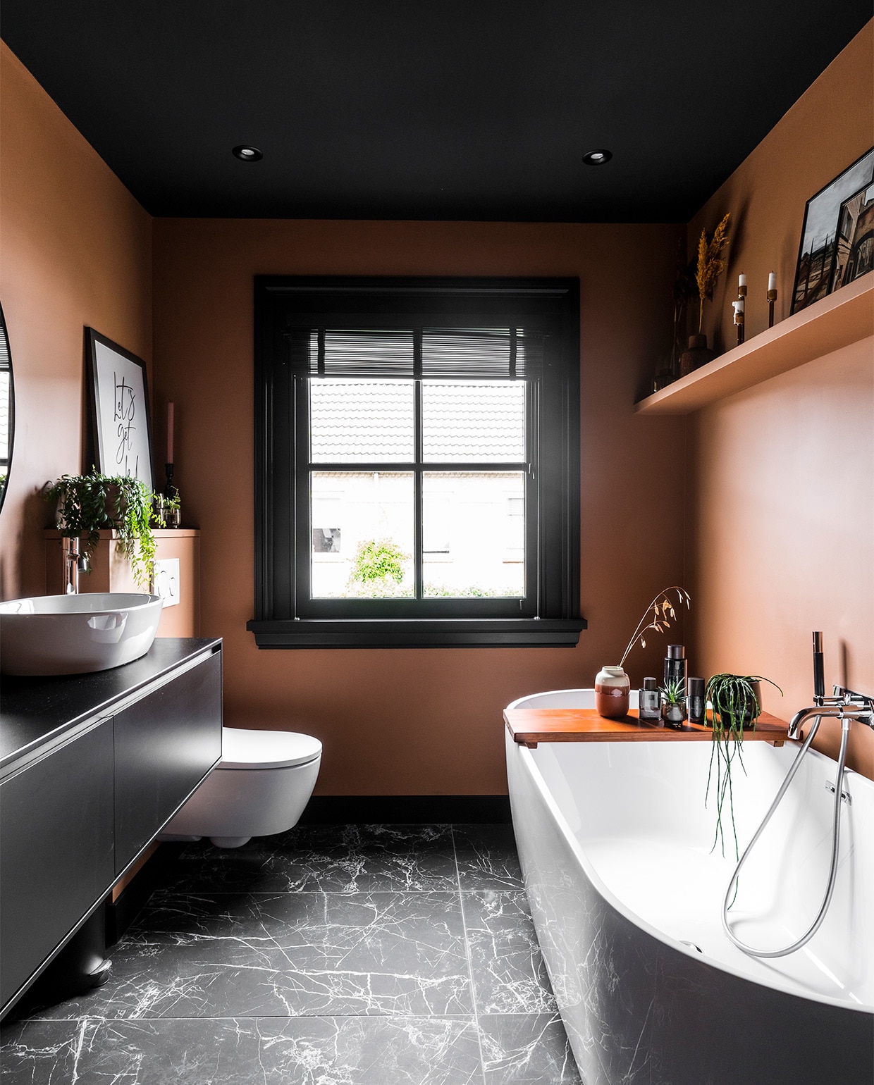 Badkamer met wandkleur Tasty Toffee Flexa, marmer look vloertegels Caesar Anima Nero mat Intercodam, bad Luca Sanitair, kraan Hotbath, badplank zelfgemaakt en in teakkleur gebeitst. Op de houten plank foto’s en souvenirs van vakantiereizen. Kozijn en raampartij zijn een renovatieingreep bedoeld voor extra licht. vtwonen binnenkijken special 2022 | producie CoCo Features | fotografie Muk van Lil, Chiela van Meerwijk