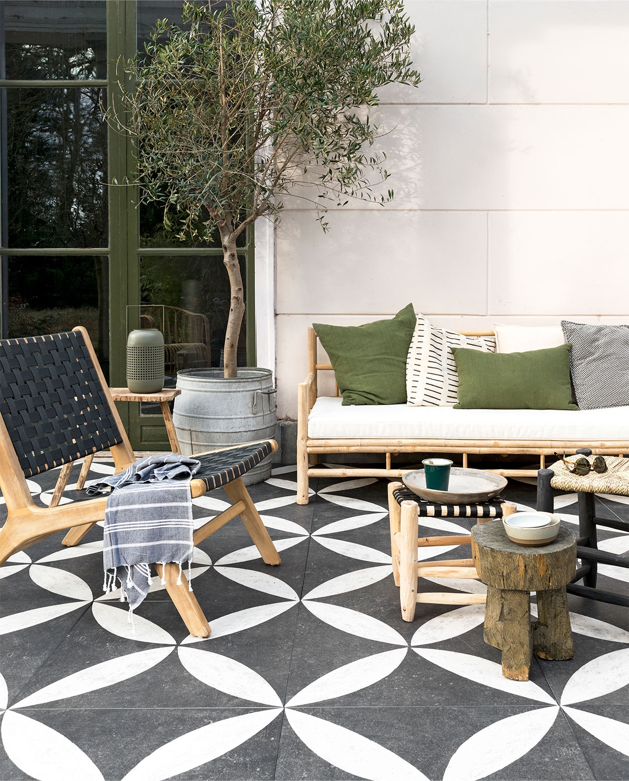 Grote zinken pot en aardewerken bord Raw Materials, krukjes met gevlochtenzitting Household Hardware, bamboe loungebank en bamboe krukje House-dressing, zonnebril, mok en schaaltjes Six and Sons, fauteuil, groene kussens, lantaarn en houten kruk vtwonen.nl, hamamdoek Ottomania, Buitentegel duostone Flower White on Black 60x60x4 cm per m² € 89,95. vtwonen 04-2018 | styling Kim van Rossenberg | fotografie Stan Koolen