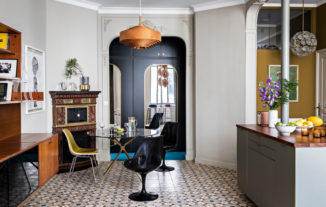 Muurverf Slipper Satin van Farrow &amp; Ball. Wandsierlijsten Orac. Vloertegels Mosaic del Sur. Teakhouten wandkast, houten hanglamp, glazen Emil Stejnar-hanglamp, eettafel en zwarte stoelen van KnollStudio vintage. , Gele stoel Eames DSX Vitra. Ingelijst kunstwerk Child van Françoise Pétrovitch. Olieverfschilderij in de keuken gevonden op de vlooienmarkt. In de op maat gemaakte wandkasten op de achtergrond zijn spiegels verwerkt. Overig onbekend. vtwonen special binnenkijken 03-2022 | productie AC Scoffoni | styling Marie-Maud Levron | fotografie Yann Deret