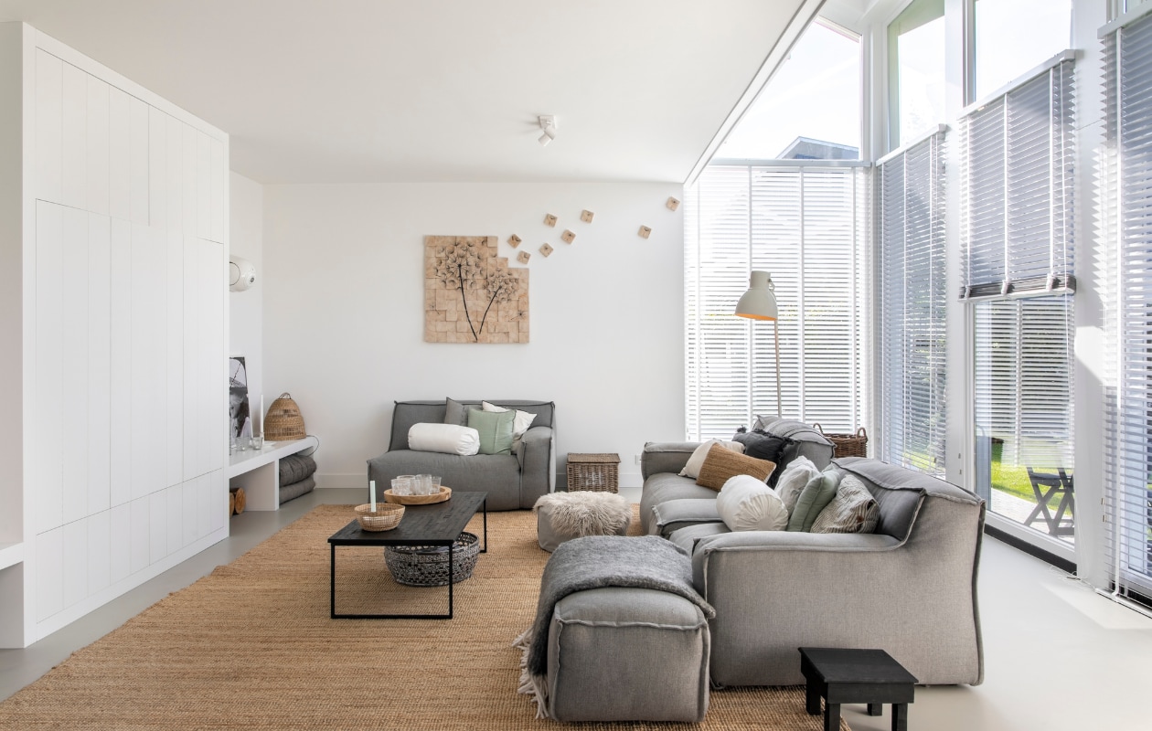 Bank De Troubadour, kunstwerk Paula Akkerman, groen kussen Maisons du Monde. Wit rolkussen, staande lamp, karpet Lohals van Ikea. Gestreept en donkergrijs kussen H&amp;M Home, naturel kussen en ronde manden Loods 5. Plafondlampen Rietveld Licht, raambekleding Joe Benelli, salontafel Ptmd, vacht Kwantum, lage vierkante bijzettafel Cooperz Wonen, plaid ’t Kethelhuys, vloer in kleur Nevada van Solo Gietvloeren, muurverf Fresh Linnen van Flexa. Overig onbekend. vtwonen 08-2022 | productie Cocofeatures | fotografie Denise Zwijnen
