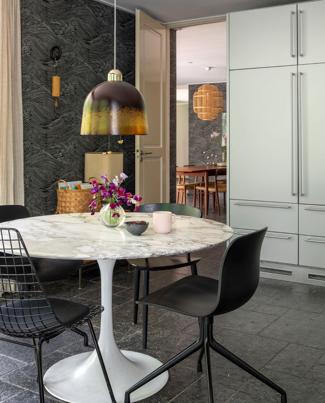 Behang Enso van Eijffinger. Marmeren vintage eettafel van Eero Saarinen, stoelen Hay en vintage, vaasje op tafel A.D. Copier van Royal Leerdam, kommetje en mok Iittala. Lamp boven tafel vintage, designer onbekend. Wandhaak Hay met Koshi gong Terra, gevlochten mand Søstrene Grene, tafellamp naast mand vintage, designer onbekend. Keuken Bulthaup, van vorige bewoners. Houten eettafel, stoelen en krukjes vintage Niels Otto Møller. Bamboe hanglamp Fine Asianliving. Vaas op houten tafel vintage, designer onbekend. Kandelaar Twiggy Squirrels via Pols Potten. NEE | vtwonen 07-2022 | productie CocoFeatures | styling Brenda van Roekel - Studio Amber Isabel en Lotje Deinum | fotografie Denise Zwijnen | tekst Saskia Borst