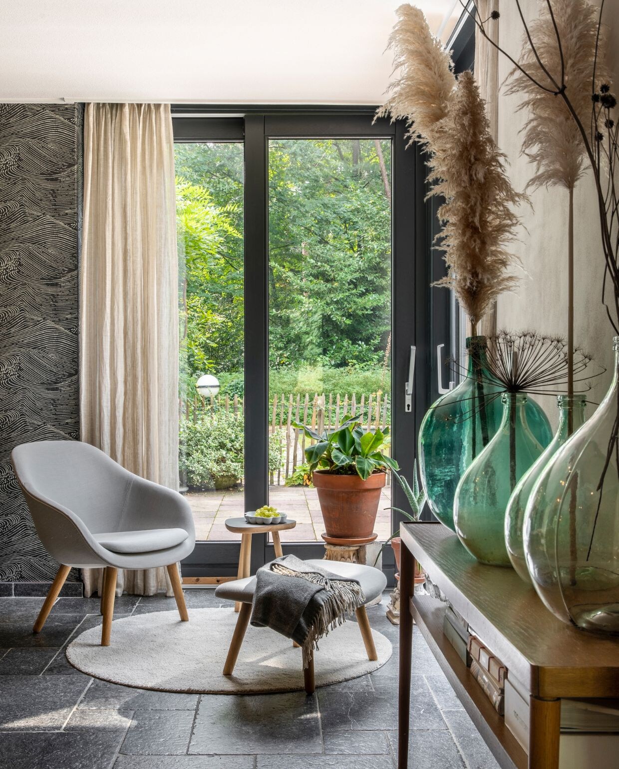 Behang Enso van Eijffinger, muurverf Elephants Breath Farrow &amp; Ball. Gordijnen Ikea. Rond vloerkleed Ikea, fauteuil met voetenbank Hay, plaid Loods 5. Bijzettafeltje designer onbekend, porseleinen schaal Bubble Dish van Studio Amber Isabel, antieke zuil met plant. Side table Ikea, gistingsflessen tweedehands, pluimen en droogbloemen uit eigen tuin. NEE | vtwonen 07-2022 | productie CocoFeatures | styling Brenda van Roekel - Studio Amber Isabel en Lotje Deinum | fotografie Denise Zwijnen | tekst Saskia Borst