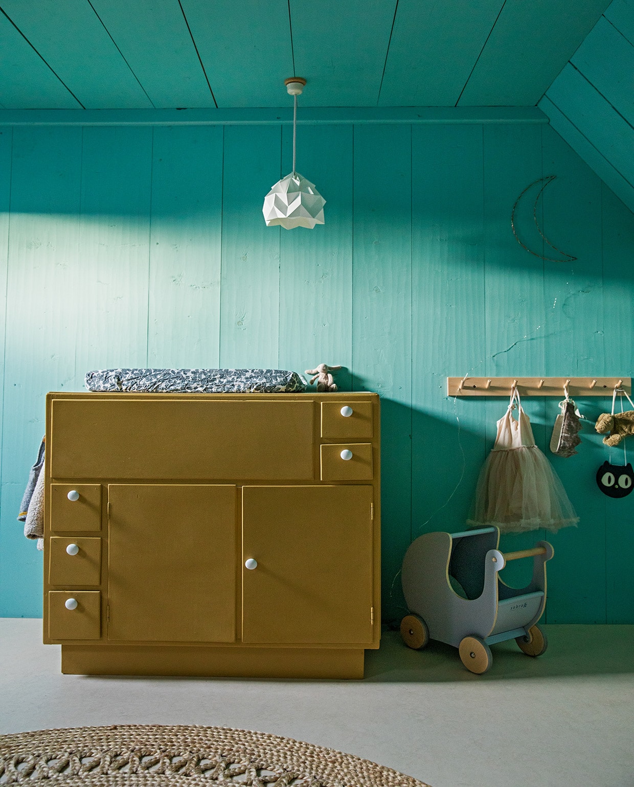Muurverf Geest van Histor. Marmoleum-vloer Forbo. Commode gemaakt door de opa van Roos’ moeder, geschilderd in okergoud van Flexa. Hanglamp Studio Snowpuppe, kapstok Iris Hantverk, commodekussen Garbo & Friends, poppenwagen antiek, vloerkleed Loods 5.