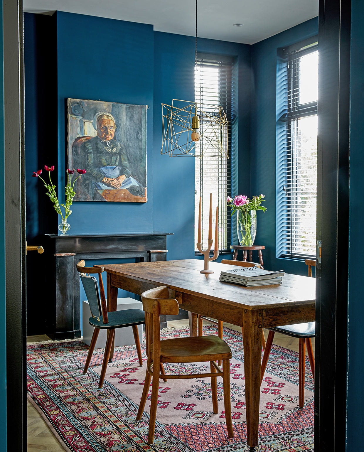 Tafel en vaas op schouw vintage, stoelen en vloerkleed kringloopwinkel. Kruk uit de inboedel van het huis, vaas op de kruk Xenos. Raambekleding Jaloezieënfabriek Zuid, muurverf Haags Blauw van Farrow &amp; Ball, schilderij Marius Richters, lamp Wattnou. vtwonen 05-2021 | fotografie Peggy Janssen | tekst Cathelijne van Wingerden\n\nvtwonen 05-2021 | eettafel van hout met kunstwerk aan de blauwe muur