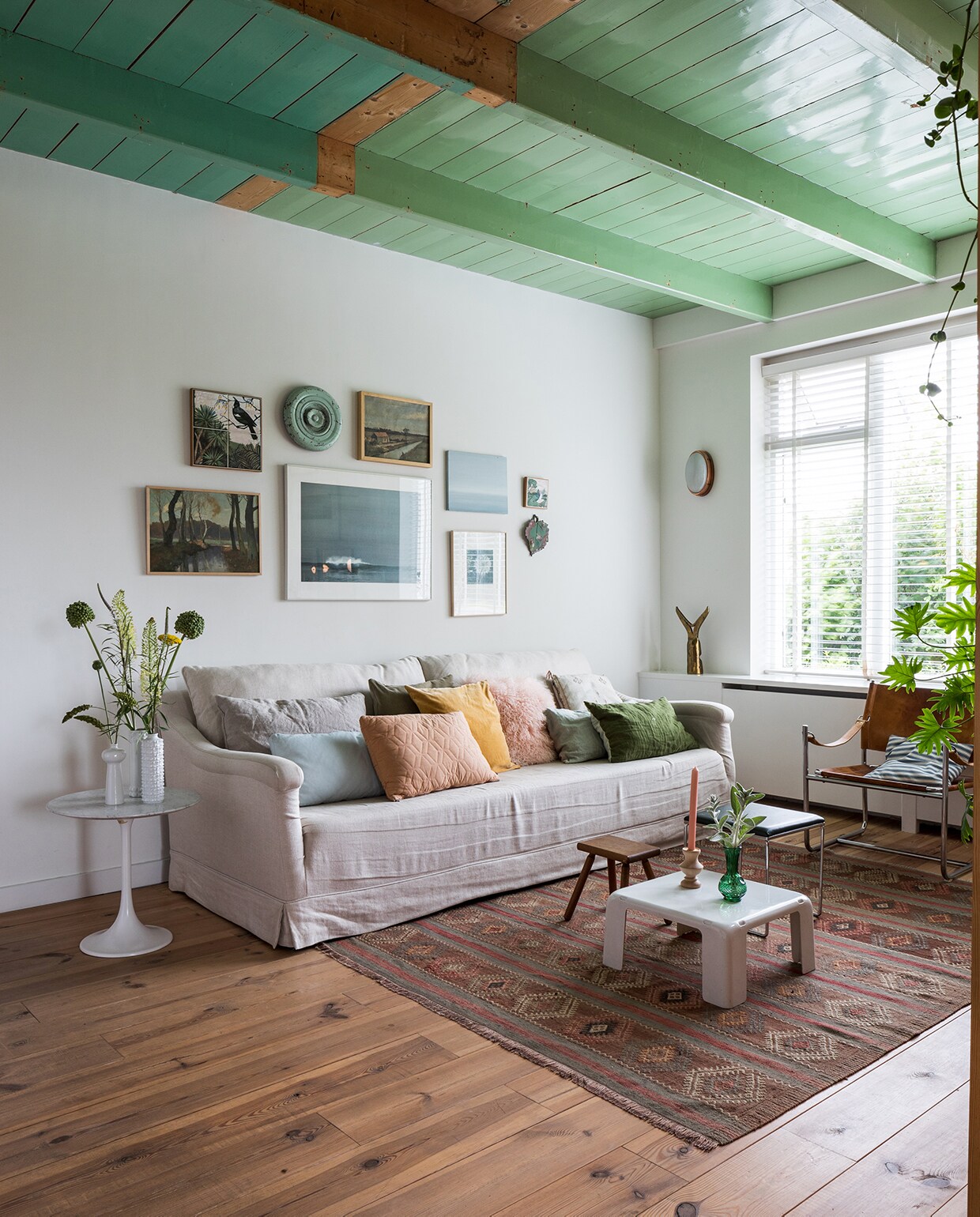 Bank Brechtje Olsthoorn Interiors, kussens zelfgemaakt, schapenvachtkussen Van Buren Bolsward. Groen linnen kussen, vazen, kandelaar, melkkruk, groene bijzettafel Sip &amp; Clara Pop-up Store. Tulp-tafel van Knoll erfstuk. Vintage kleed Richard Houpt Noordermarkt Amsterdam. Witte vintage bijzettafel B&amp;B Italia. Vintage buisframe stoel en wandlamp Marktplaats. Herkomst bronzen voorwerp onbekend. Aan de muur met de klok mee: tegel vogel uit Nieuw-Zeeland, groene deksel van ouderwets toilet, schilderij landschap rommelmarkt, horizonschilderij Auke van der Meulen, tegel uit Nieuw-Zeeland, aardewerken blad Trees In der Rieden, zeefdruk paardenbloem Ray Daal, grote foto cadeau gekregen, schilderij bomen kringloop. vtwonen DIY special 01 2020 | landelijke woonkamer met lijsten wand op houten vloer binnenkijken Hemelum