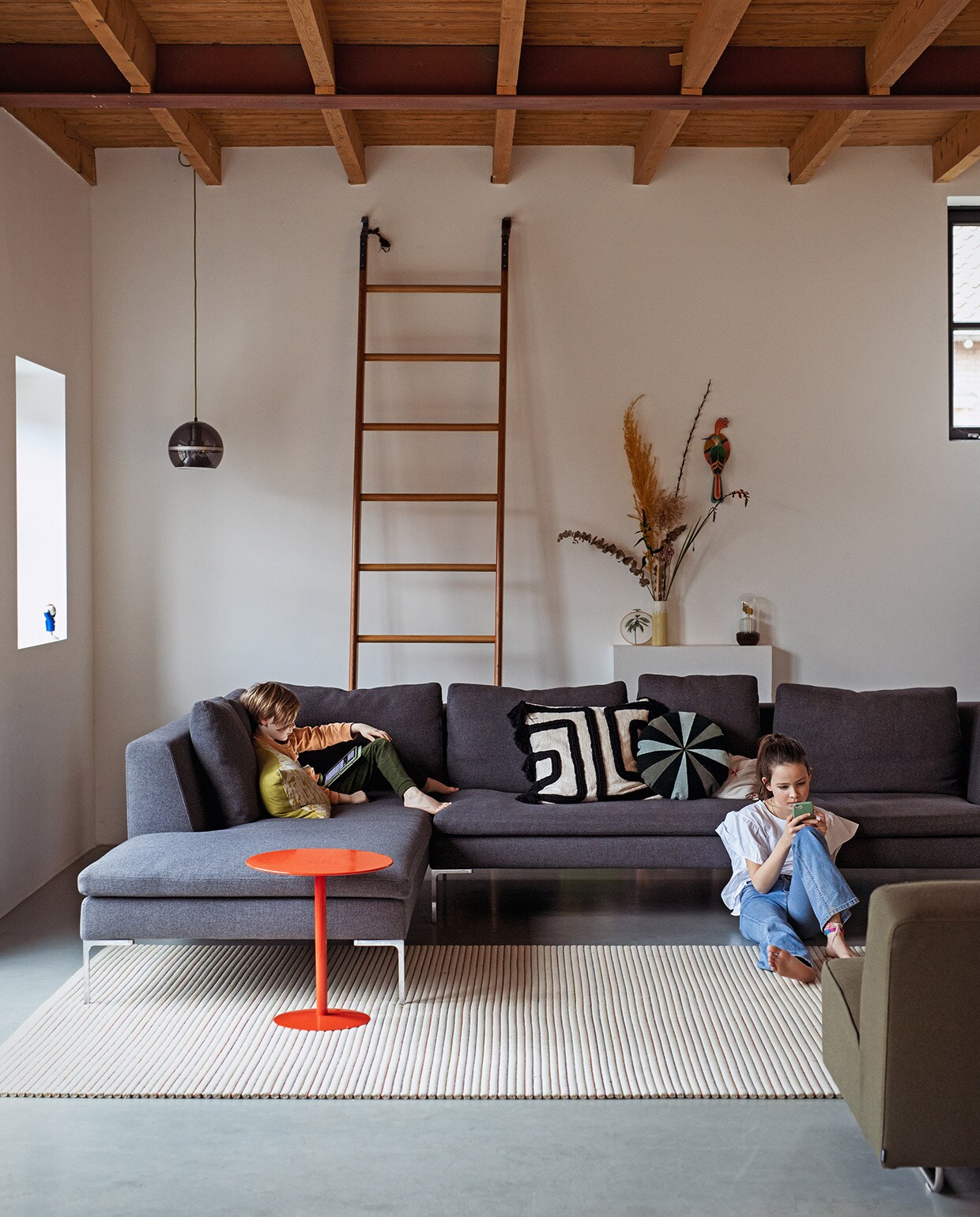 Bank Brechtje Olsthoorn Interiors, kussens zelfgemaakt, schapenvachtkussen Van Buren Bolsward. Groen linnen kussen, vazen, kandelaar, melkkruk, groene bijzettafel Sip &amp; Clara Pop-up Store. Tulp-tafel van Knoll erfstuk. Vintage kleed Richard Houpt Noordermarkt Amsterdam. Witte vintage bijzettafel B&amp;B Italia. Vintage buisframe stoel en wandlamp Marktplaats. Herkomst bronzen voorwerp onbekend. Aan de muur met de klok mee: tegel vogel uit Nieuw-Zeeland, groene deksel van ouderwets toilet, schilderij landschap rommelmarkt, horizonschilderij Auke van der Meulen, tegel uit Nieuw-Zeeland, aardewerken blad Trees In der Rieden, zeefdruk paardenbloem Ray Daal, grote foto cadeau gekregen, schilderij bomen kringloop. vtwonen 03-2021 | Styling Ilona Jongepier | Fotografie Dana van Leeuwen | Tekst Sara Madou\nvtwonen 03-2021 | grijze hoekbank in woonkamer ladder tegen muur