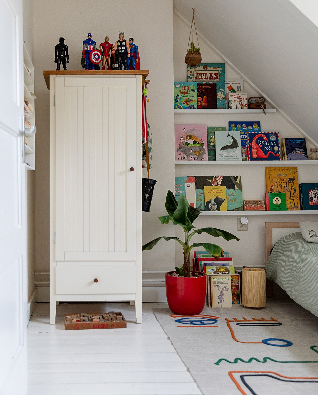 Witte kast, rode pot, houten letterdoos, letterbak aan de wand en Marokkaanse lamp op de grond kringloop. Tapijt Kwantum. Verzameling superhelden van de rommelmarkt. Bananenplant Intratuin. Bed en beddengoed Ikea. Grijs kussen met vogels Zilalila. Vintage verrekijker van opa. JA | vtwonen 13-2021 | styling Ilona Jongepier | fotografie Dana van Leeuwen