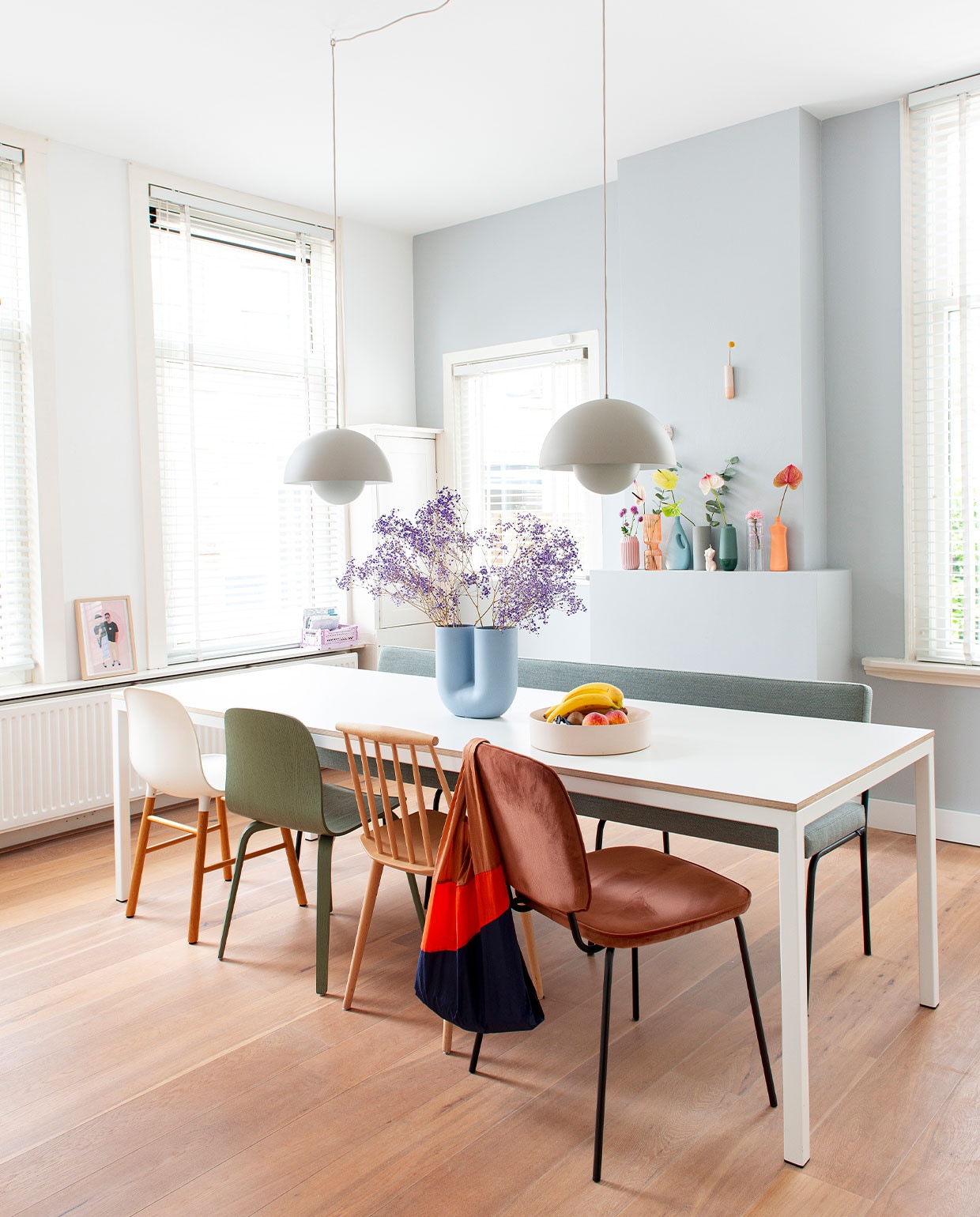 Muurverf Wild Dove van Flexa. Hanglampen FlowerPot van Verner Panton via &amp;Tradition. Tafel op maat gemaakt. Vaas Kink Muuto. Fruitschaal Fest Amsterdam. Witte stoel Normann Copenhagen. Groene stoel Muuto via Femkeido. Spijlenstoel Hay. Oranje stoel House Doctor. Bank Monday Fest Amsterdam via Wonenmetlef. Illustratie op vensterbank Melissa van Roon, MVR Design. Vazen op schoorsteen: roze en oranje Loods 5, blauwe en oranje fles Foekje Fleur, grijs Ferm Living, groen Hella Duijs. Wandvaas Studio Harm &amp; Elke. NEE | vtwonen binnenkijken special balans 12-2021 | styling Jeske Weel | fotografie Caroline Coehorst | tekst Chris Muyres