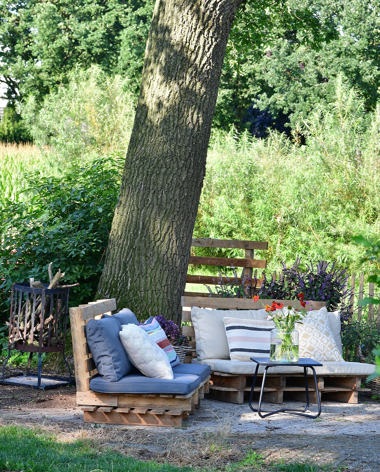 Een fijne zitplek op warme dagen: de zelfgemaakte palletbanken onder de oude eikenboom. Op de achtergrond wilgenbomen. Matraskussens en tafel Kwantum, sierkussens van onder andere fairtrade label Nic &amp; Mic (streep links). NEE | vtwonen TUIN special 5 | fotografie Sietske de Vries | tekst Monique van der Pauw