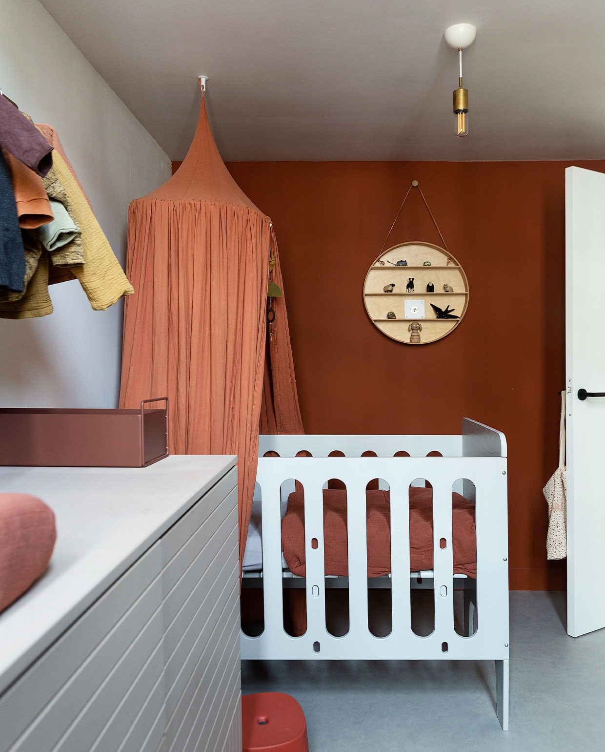 Babybed Timmer Toko van Overdijk, hemel Nobodinoz, beddengoed en aankleedkussen Garbo &amp; Friends. Rond wandobject met spullen van Mini &amp; Momo. Opstapkruk AliExpress, tas aan deurklink Archive Store. Commode eigen ontwerp op basis van keukenkasten Ikea, lijnen zelf erin gefreesd. Metalen bak Ferm Living. Plafondlamp Watt. Vloer Marmoleum van Forbo. Muurverf Pure Sienna van Flexa. vtwonen 06-2022 | productie Martine Benjamins | styling Emmy van Dantzig | fotografie Jansje Klazinga