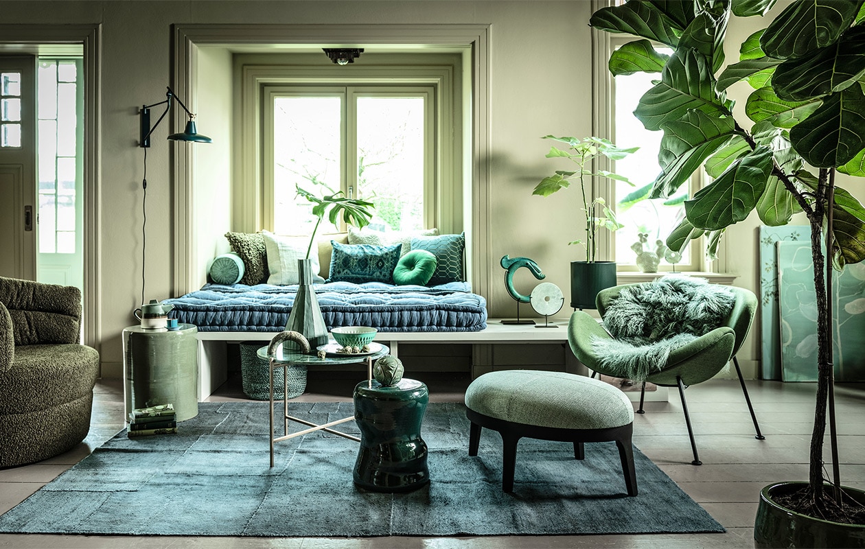 Fauteuil links (flinders.nl). Bank op maat gemaakt (com-anders.nl). Zitting bank twee stuks op maat gemaakt, 210×80 cm, geel/groen langwerpig kussen (coco-mat.com). Fauteuil Orange Slice van Artifort, cactus van Hay (homestede.nl). Marmeren tafel, kruk links, kruk midden, kussen langwerpig met oude opdruk, ronde sierbol op kruk prijs op aanvraag, sierobject draak (loods5.nl). Poef, grof kussen, kussen geometrisch (covanderhorst.nl). Marmeren sierobject op tafel (wonenmetlef.nl). Bord, per 6 (livingandcompany.com). Theepot (bijenkorf.nl). Vloerkleed, 242×178 cm, langwerpig kussen achter, lichtgroen kussen voor, mand (couleurlocale.eu). Wandlamp Hector van Dutchbone, rond kussen Mandarin van Kaat Amsterdam, Ø 40 cm (vtwonen.nl). Notitieboeken (maisonnl.com). Espressokop en schotel schaal (ottomania.nl). Rolkussen (hinck.nl). Hoge vaas, ronde siervorm op bank en in vensterbank (badenbaden.nl). Schilderijen achter Branch on a green canvas, 80×100 cm, en Floral sketch, original painting, 70×80 cm prijs op aanvraag (maartjevandennoort.nl). Plantenpot op pootjes (fermliving.com). Plantenpot op grond uncwardrobe.com). JA | vtwonen 04-2020 | styling Liza Wassenaar | fotografie Sjoerd Eickmans