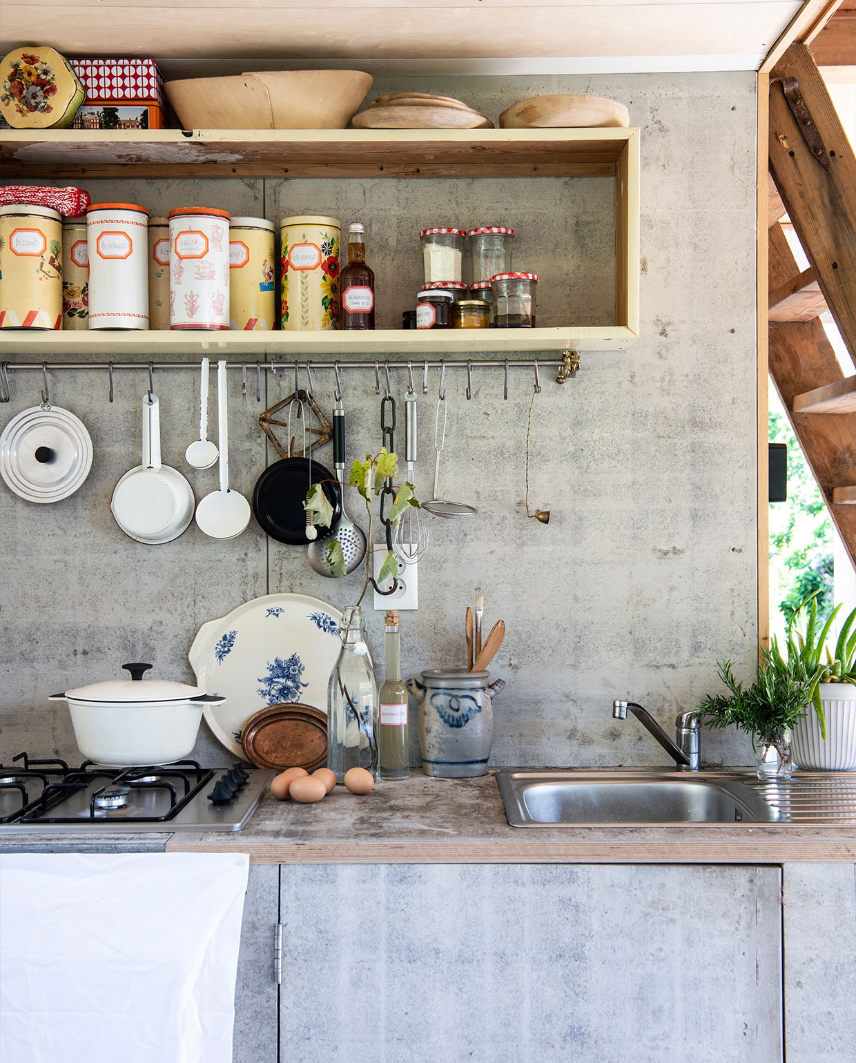 Keuken door Martin zelf gebouwd van steenschotten, waar ook het terras rondom het tuinhuis van gemaakt is. Witte braadpan Ikea, bord met blauwe bloemen erfstuk, Keulse pot vintage, railing met haken Ikea, houten borden HKliving, vintage beschuitbussen als voorraadblikken, lepels aan railing Van Manen aan Tafel, witte steelpan Dille &amp; Kamille. vtwonen tuin special 5 | Productie Insidehomepage | Styling Myla Griese | Fotografie Louis Lemaire