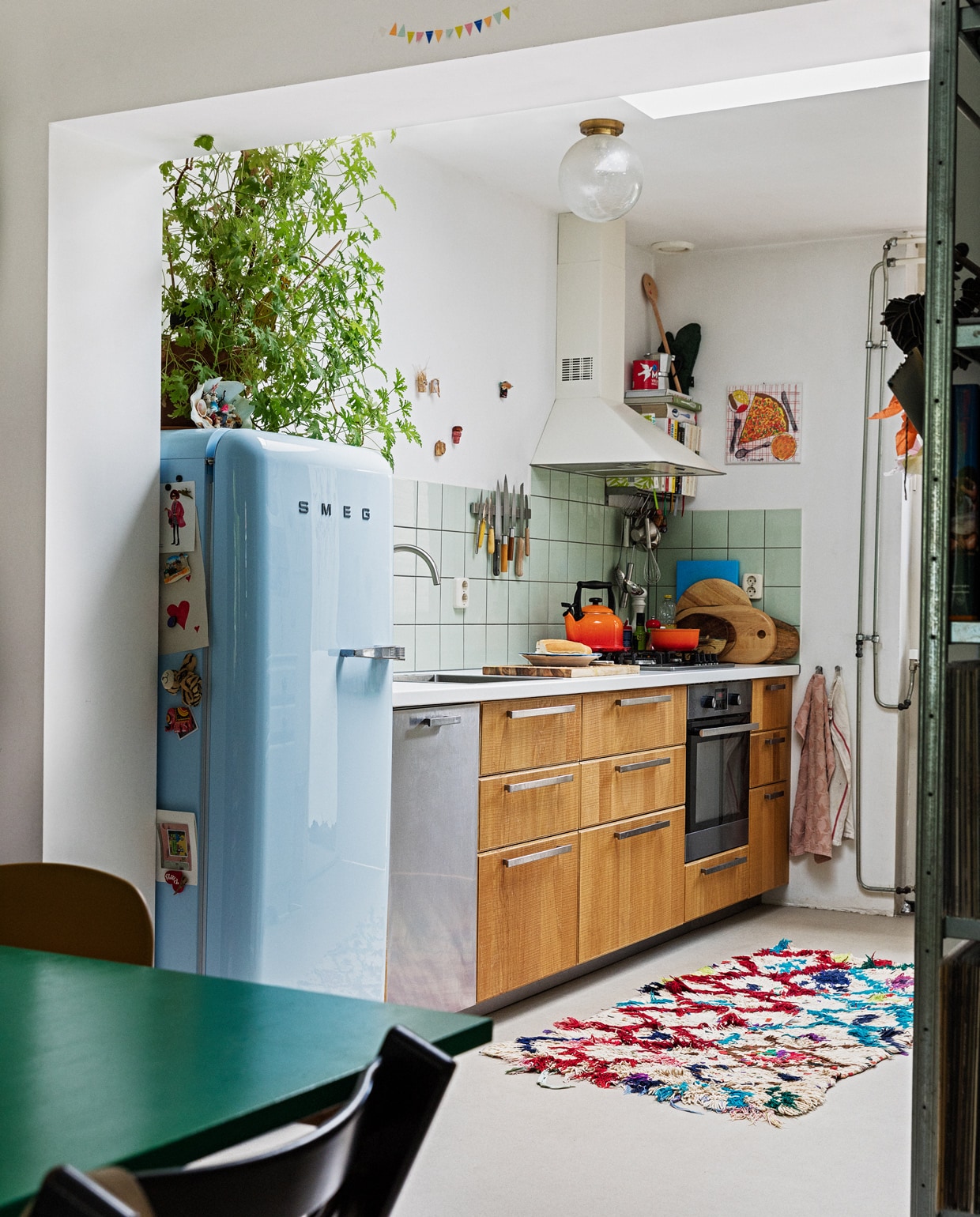 Koelkast Smeg. Geraniumplant zelf gestekt. Kitsch schelp op koelkast souvenir uit Griekenland. Magneet-souvenirs meegenomen van verschillende reizen. Fluitketel en pan Le Creuset. Messen Suncraft, Opinel, Dille &amp; Kamille, Pallarès Solsona. Snijplanken Hay, Ikea, Serax. Soeplepel Brabantia. Rvs keukengerei Dille &amp; Kamille. Schaaltje van een pottenbakkersmarkt. Afzuigkap en oven Ikea. Ovenhandschoen Kook. Pollepel Kookhuis aan de Maes. Handdoeken Sissy-Boy. Tapijt The Souks. Pizza-schilderij Oskar. Ronde plafondlamp lampenwinkel.nl. vtwonen 13-2021 | styling Ilona Jongepier | fotografie Dana van Leeuwen