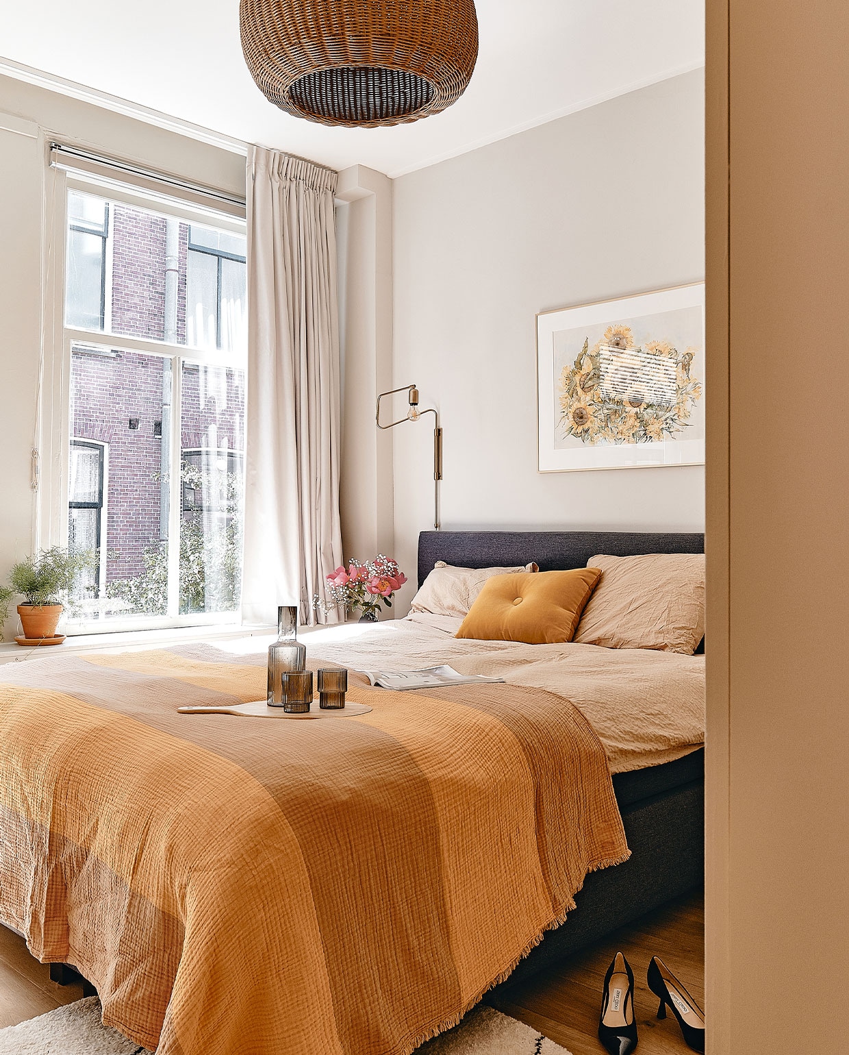Muurverf Ammonite van Farrow &amp; Ball. Wandlamp Swing van House Doctor. Hanglamp Braided Belly van Ferm Living. Ingelijste zonnebloemen van Maartens ouders. Beddengoed Crisp Sheets, oranje kussen en sprei Hay. Dienblad Ikea, karaf en glazen Ferm Living. Vloerkleed Benuta, pumps Jimmy Choo. Kast Pax van Ikea, geverfd in Hay van Farrow &amp; Ball. vtwonen 05-2022 | productie Cocofeatures | fotografie Roos Oosterbroek