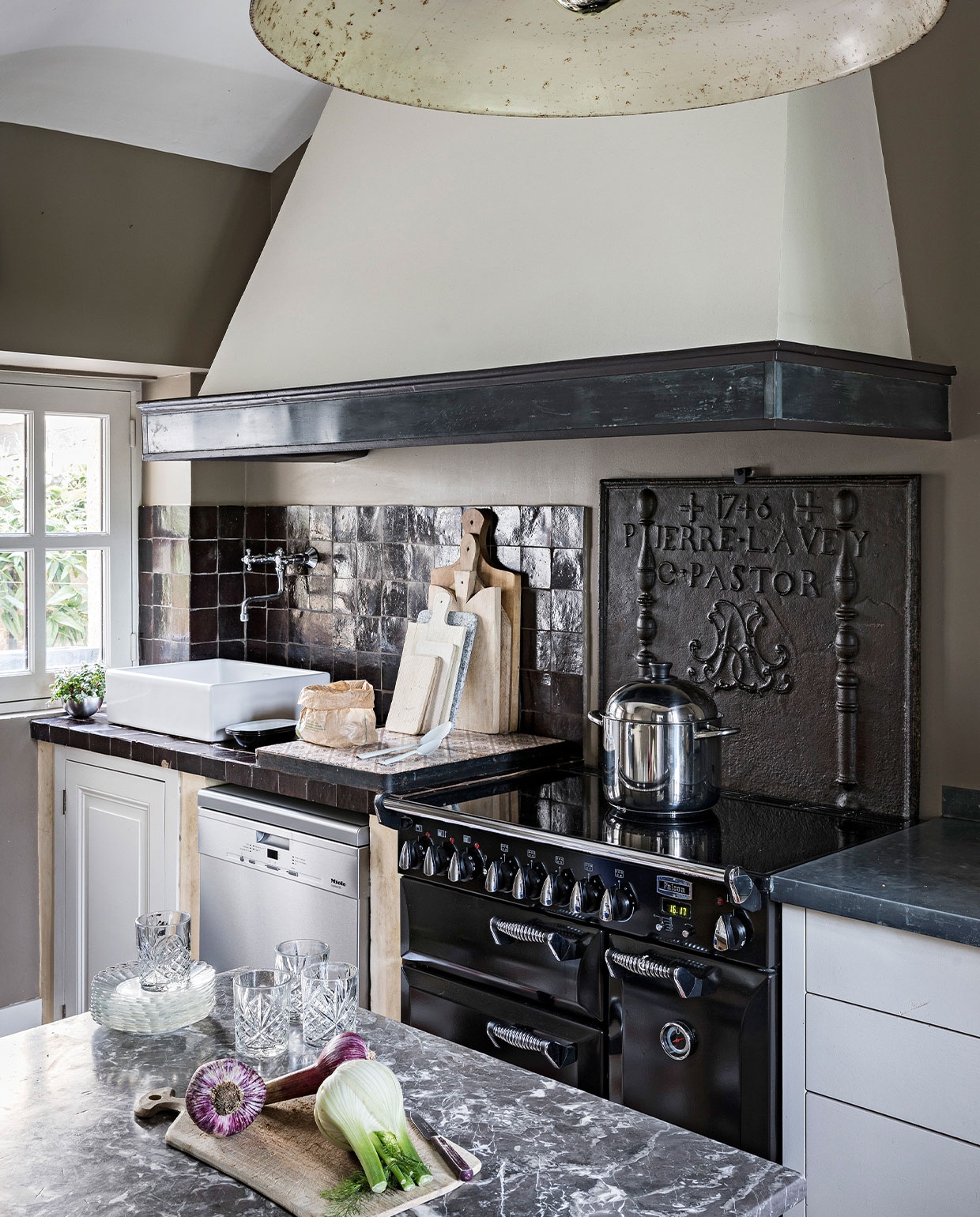 Wandtegels zelliges van Emery &amp; Cie, boven het fornuis een 13de-eeuwse gietijzeren achterplaat gevonden bij een schroothandel in Sartre, aan het plafond een vintage stallamp. NEE | vtwonen 4-2022 |  styling  Marie-Maud Levron | fotografie Yann Deret | tekst Anne-Catherine Scoffoni | tekstbewerking Chris Muyres