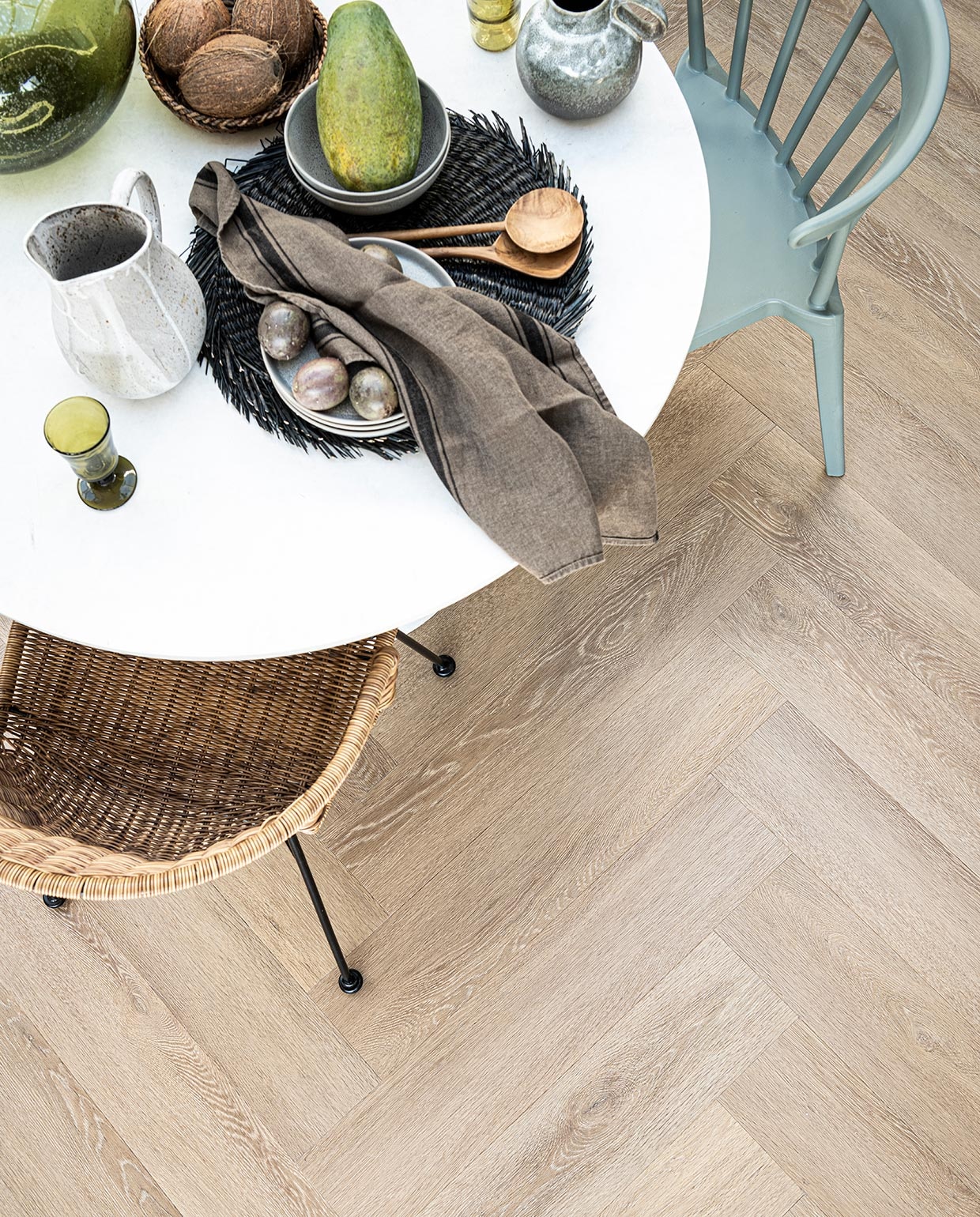 Pvc-vloer Herringbone Sun Kissed, 76,5×15,3 cm, click 75×15 cm M.m.v. basiclabel.nl (eettafel en eetkamerstoelen van Woood), vtwonen.nl (grijze borden, kommen, placemats, kruik met handvat, alles van Bloomingville), sissy-boy.com (grijze kan wit geverfd), hm.com (mand), paviljoennoord.com (groen wijnglas, groene waterglazen, houten bestek, groene glazen vaas, theedoek). Overig privébezit. vtwonen 06-2021 | productie Tanja Saarberg | styling Danielle Verheul | fotografie  Sjoerd Eickmans