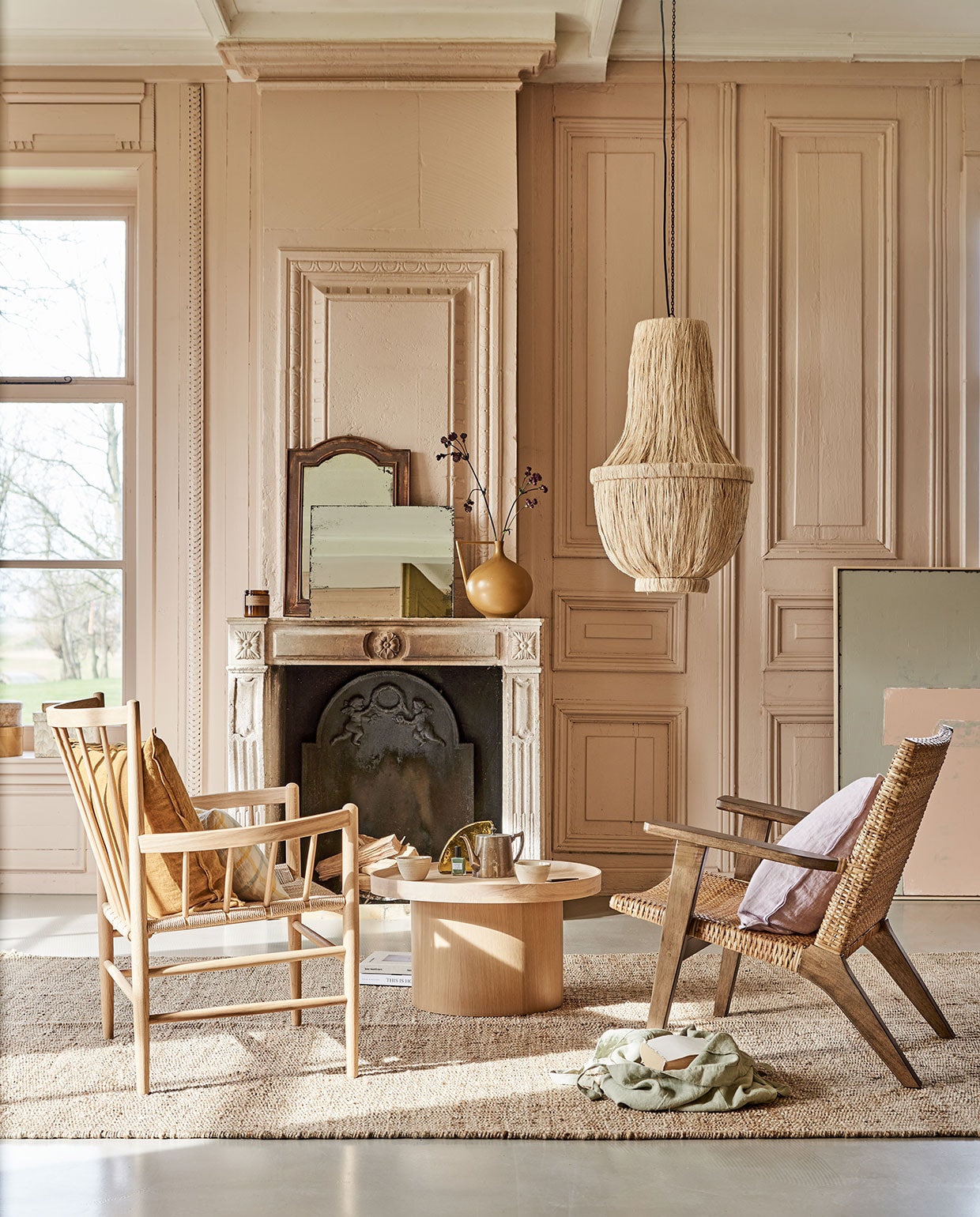 Roze tinten aan de muur met neutralen vloerkleed en schouw | Fauteuil links FD Møbler J82 Jørgen Baekmark Lounge Chai,, linnen tas, geruit kussen, geurkaars, mokken op tafel (letoile.store). Fauteuil rechts, vloerkleed, 200×300 cm, kunstwerk Abstract 100×120 cm, rechthoekige spiegel antieklook, vaas in mosterdkleur, glazen artwork op tafel (hkliving.nl). Zacht lila kussen van Bungalow, okergeel kussen, papieren dozen (scandicliving.nl). Oude spiegel met houten rand (com-anders.nl). Salontafel Plateau van Bolia, 34,3×61 cm (hxØ) (vtwonen.nl). Kroonluchter van gedroogde bananenbladeren (rivieramaison.com). Kleur op de wanden vergelijkbaar met Setting Plaster No.231 van Farrow &amp; Ball. vtwonen 05-2021 | styling Marlies Does | fotografie Bart Brussee