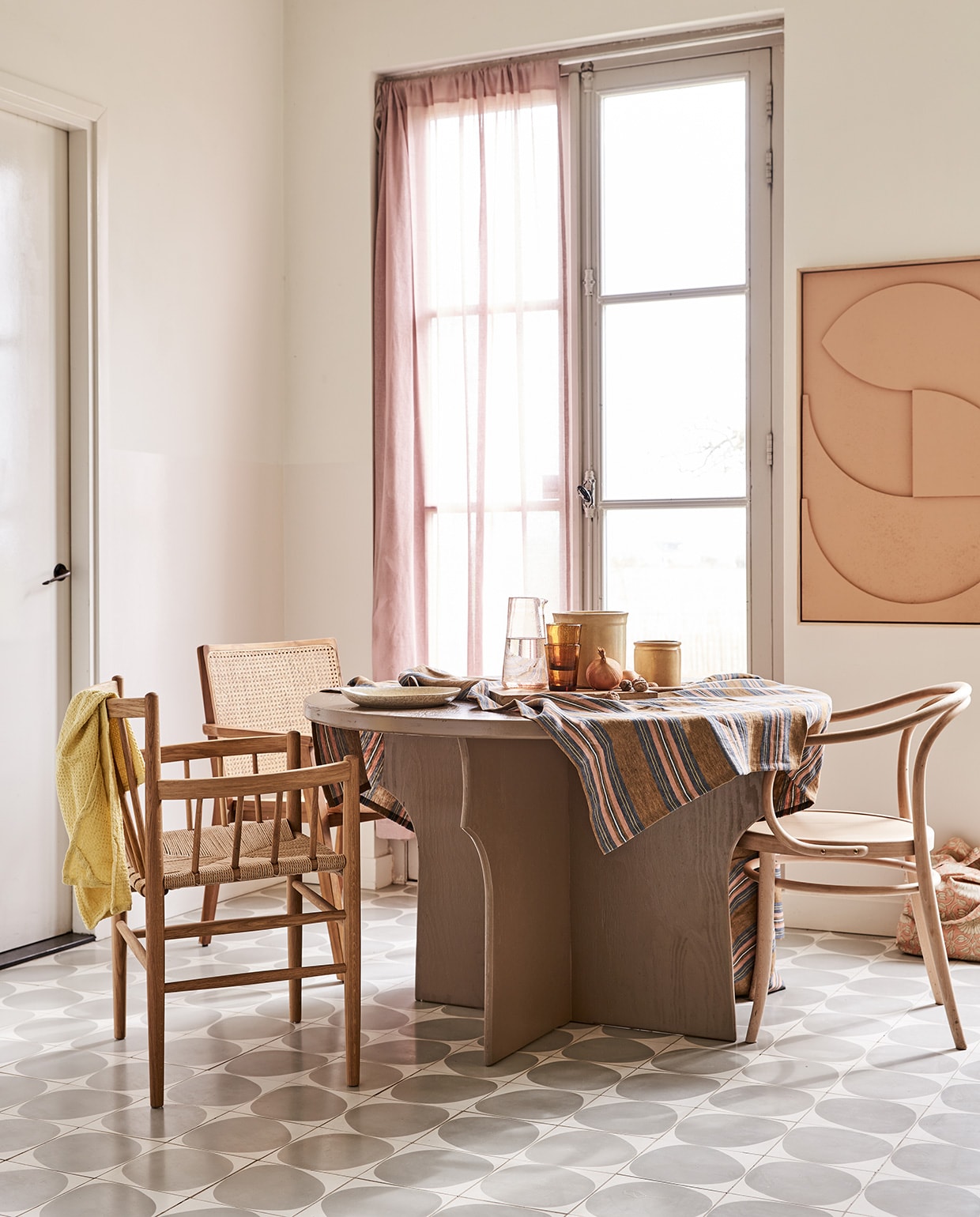 Tafel gemaakt naar eigen ontwerp en geschilderd in kleur Mexican Sand, prijs op aanvraag (com-anders.nl). Stoel met spijlen FD Møbler J81 Jørgen Baekmark (letoile.store). Stoel met webbing zitting Nova (vtwonen.nl). Stoel Thonet-model (dille-kamille.nl). Tafelkleed (Libeco). Karaf, water- glazen Salon Amber, bord, theedoek wafel in kleur Lemon, tas met roze bloemen (scandicliving.nl). Artwork aan de wand in kleur perzik, 60×80 cm (hkliving.nl). Gordijn van voile stof Cosia in de kleur Orchid van Designers Guild (wva.nl). vtwonen 05-2021 | styling Marlies Does | fotografie Bart Brussee