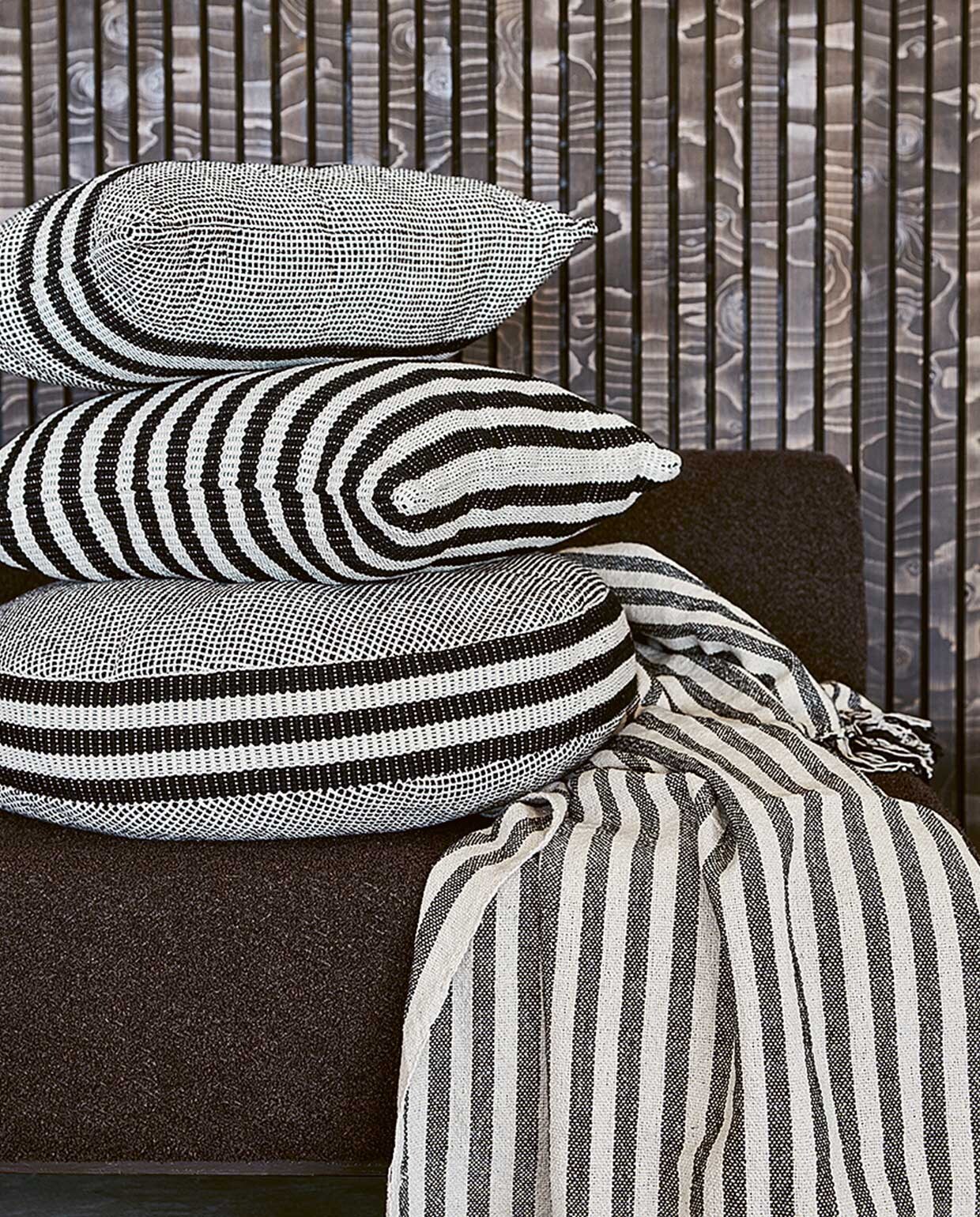 Zwart-witte kussens van chambray: rechthoekig 60×35 cm € 24,95, rond 60×10 cm (Øxh) € 39,95, vierkant 50×50 cm € 29,95. Zwart-wit gestreepte plaid van chambray met geknoopte franjes, 130×170 cm € 49,95. vtwonen 04-2022| Productie Tanja Saarberg | Styling Marlies Does | Fotografie Bart Brussee