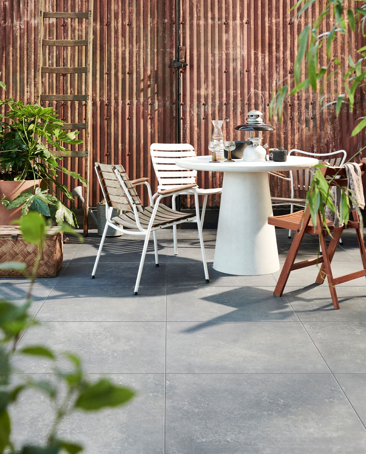 Buitentegels Solostone Uni Chateau Black, 70x70x3,2 cm per m² € 69,95.M.m.v. basiclabel.nl (betonlook tafel van Damon), vtwonen.nl (witte metalen stoel van Zuiver), com-anders.nl (oude ladder, houten kist, oude gieter, klapstoel, lantaarn, rieten mand), deens.nl (bloempot met onderstel van Ferm Living, stof over stoelleuning, kan, glazen), vandijkenko.nl (stoelen van ijzer en hout). vtwonen 04-2022 | Productie Tanja saarberg | Styling Marlies Does | Fotografie Sjoerd Eickmans\n