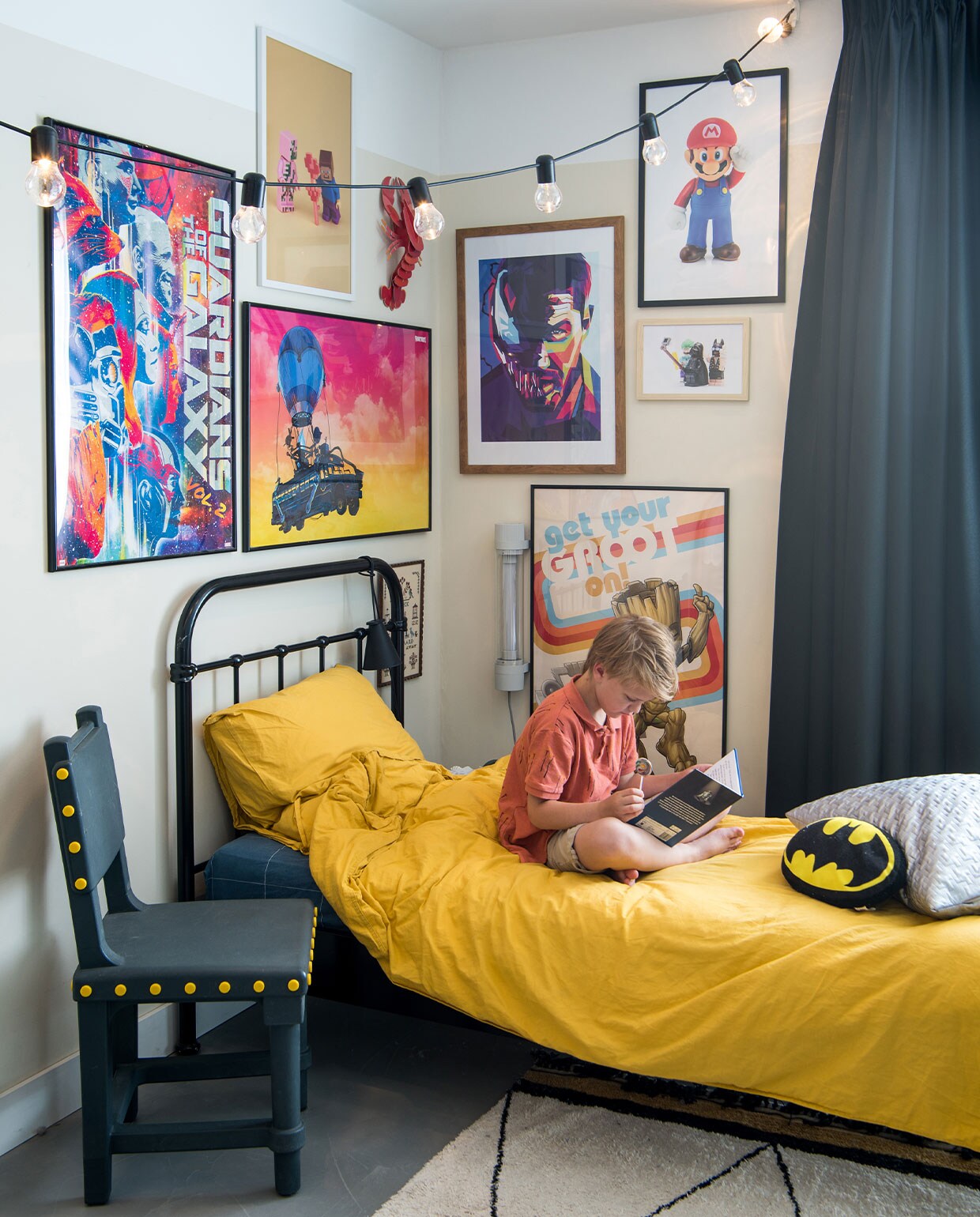 Stoel Studio Job, lichtsnoer Ikea, posterwand is combinatie van gekochte en zelf op maat gemaakte posters, kreeft Studio Roof, lamp aan bed Menu, lamp aan muur Fatboy, dekbedovertrekset H&amp;M, vloerkleed onbekend, Batman-kussen Kruidvat, zilveren kussen zelfgemaakt. Muurkleur RAL 1013, zwarte overgordijnen op maat gemaakt door stoffeerder. vtwonen 2-2022 | styling Kim de Groot | fotografie Anke Leunissen | tekst Mirjam Rosema-Verhulst